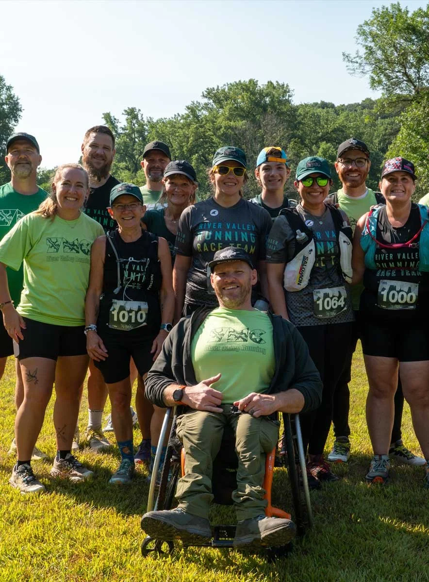 Group of runners, including a man in a wheelchair, posing outdoors on a grassy field with trees in the background during a race event.