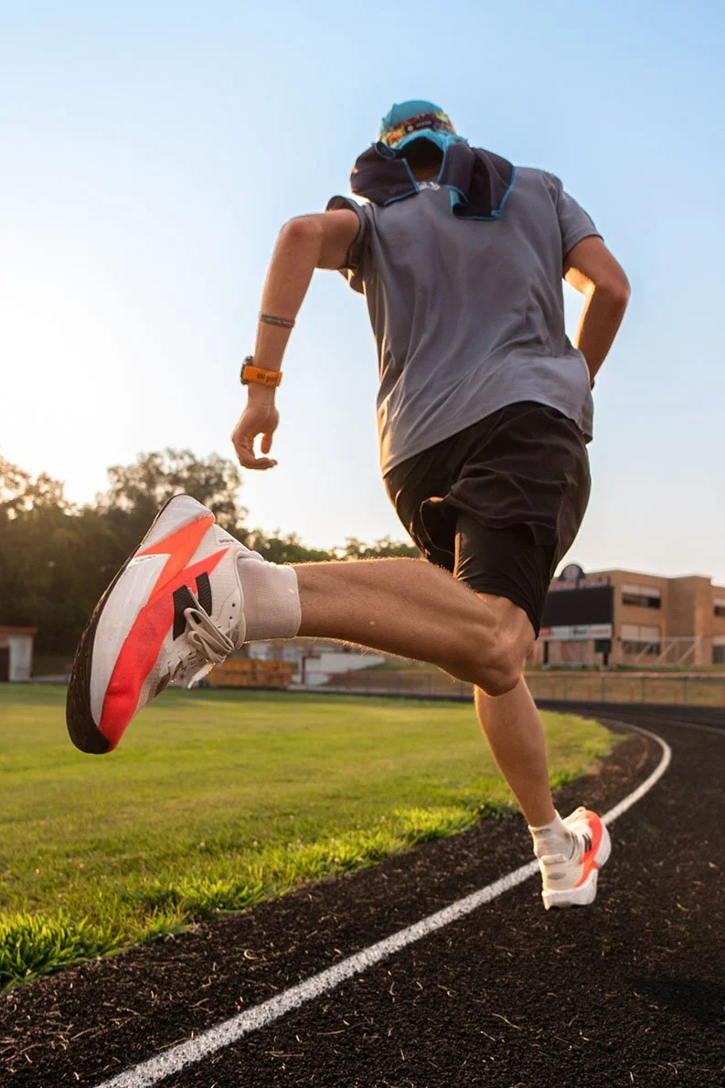 A person running on a track field during sunset, wearing a gray shirt, black shorts, white and red running shoes, with a towel or shirt draped over their head.