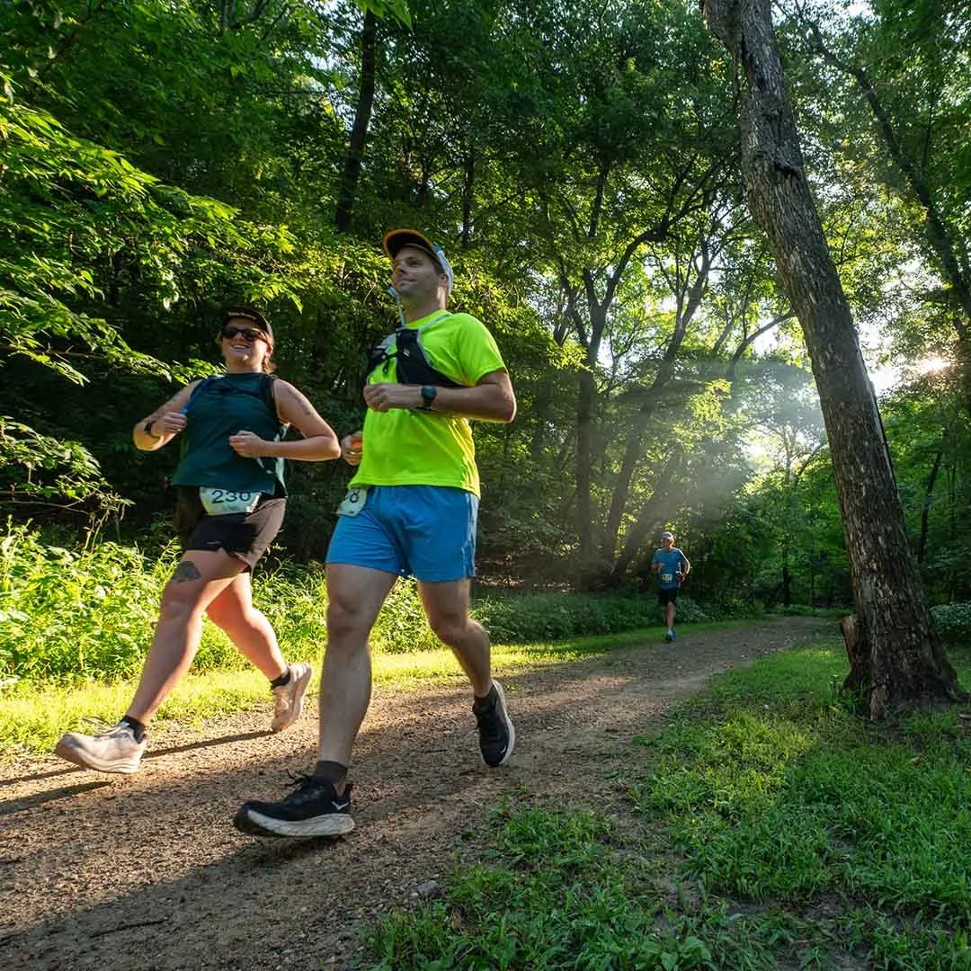 Group of people running on a forest trail surrounded by tall trees and green foliage during daylight.