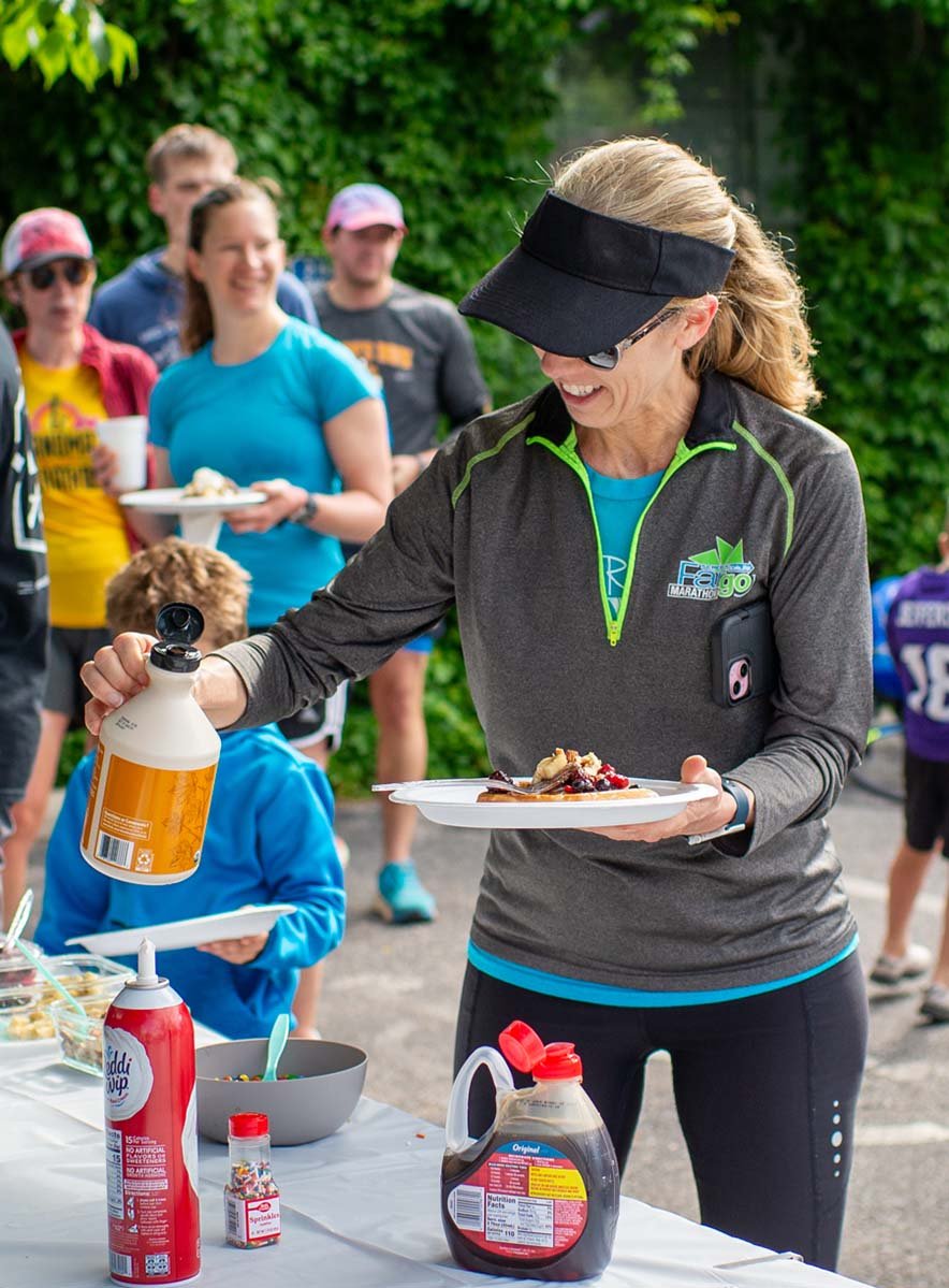 A woman in athletic clothing and a visor is pouring syrup on a plate of dessert at an outdoor gathering, with a group of people standing behind her, some holding plates and drinks, in a setting with green foliage.