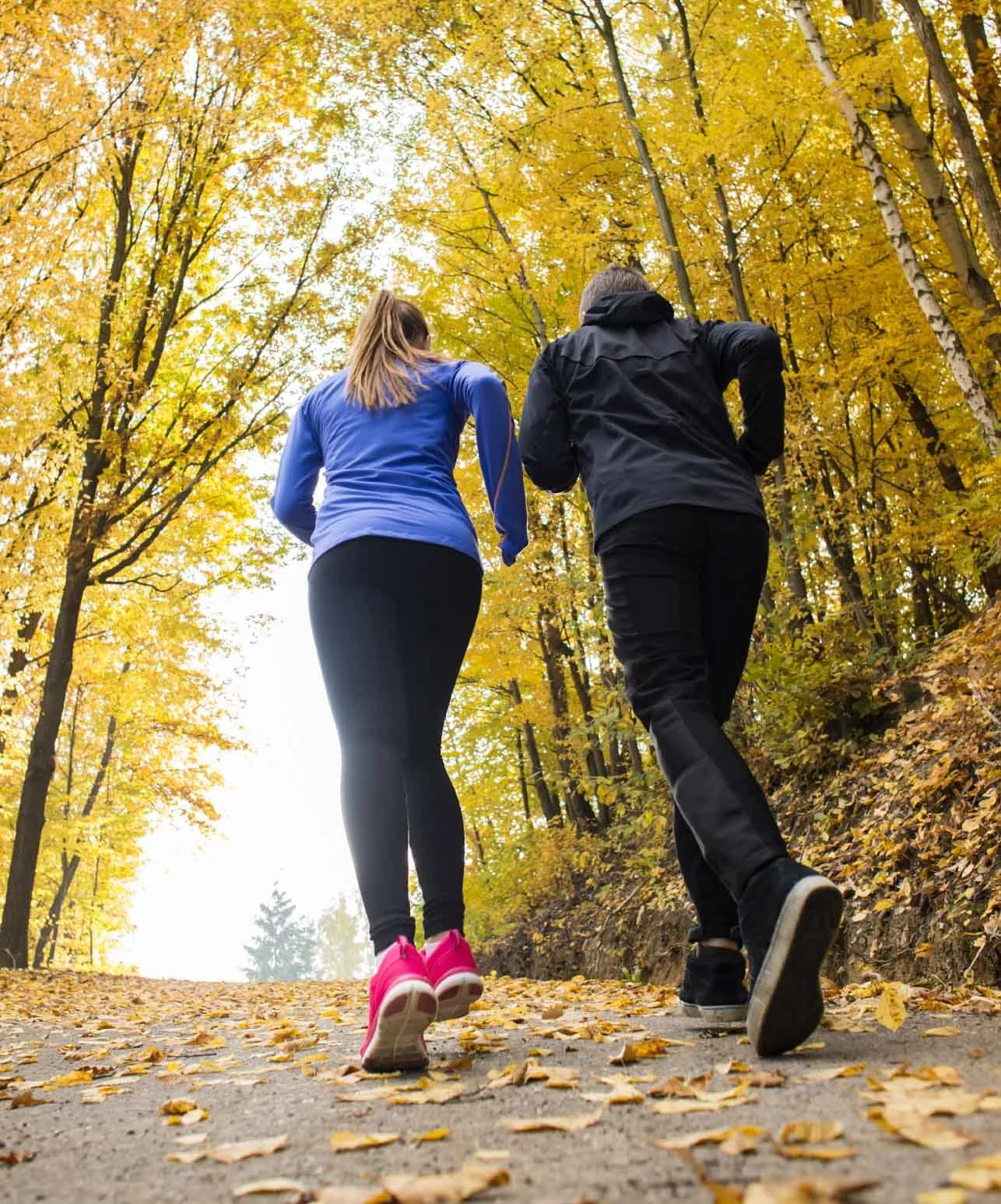 A man and a woman are running on a leaf-covered trail surrounded by colorful autumn trees, viewed from behind.