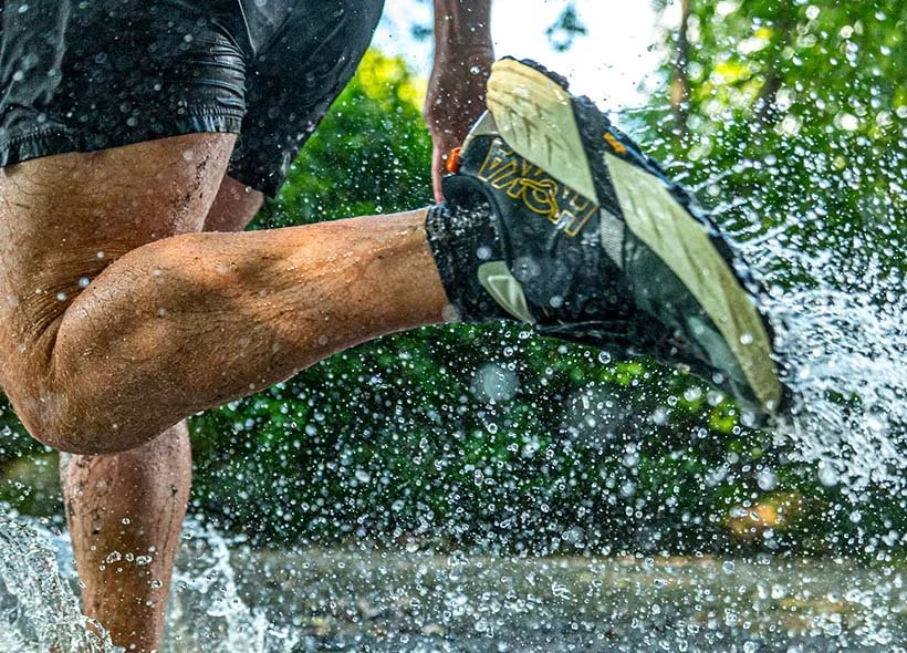 Close-up of a person running in water outdoors, wearing black shorts and water shoes, with water splashing around.