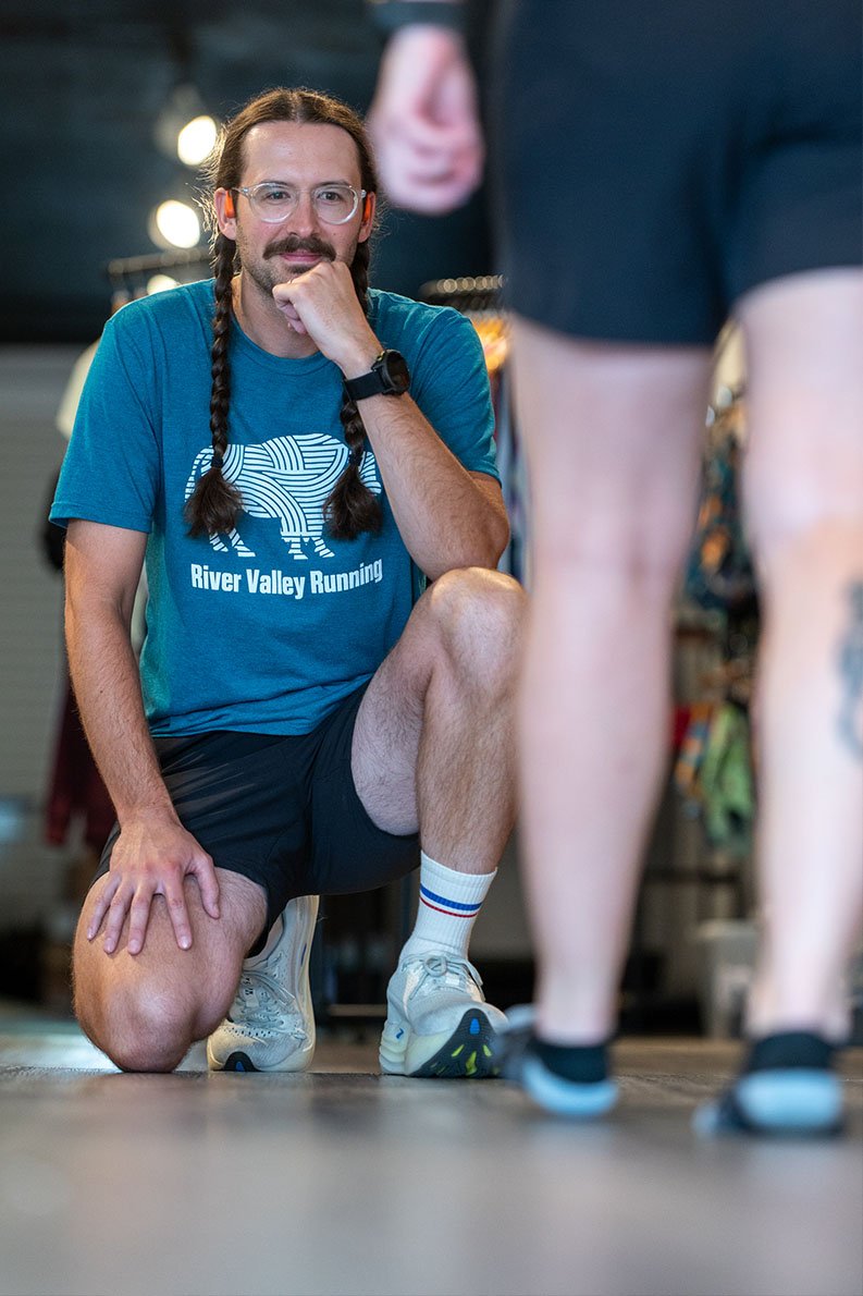 A man with long braided hair and glasses kneeling on one knee in a gym, wearing a blue T-shirt with "River Valley Running" written on it, and looking at a person in front of him who is wearing athletic shorts and sneakers.