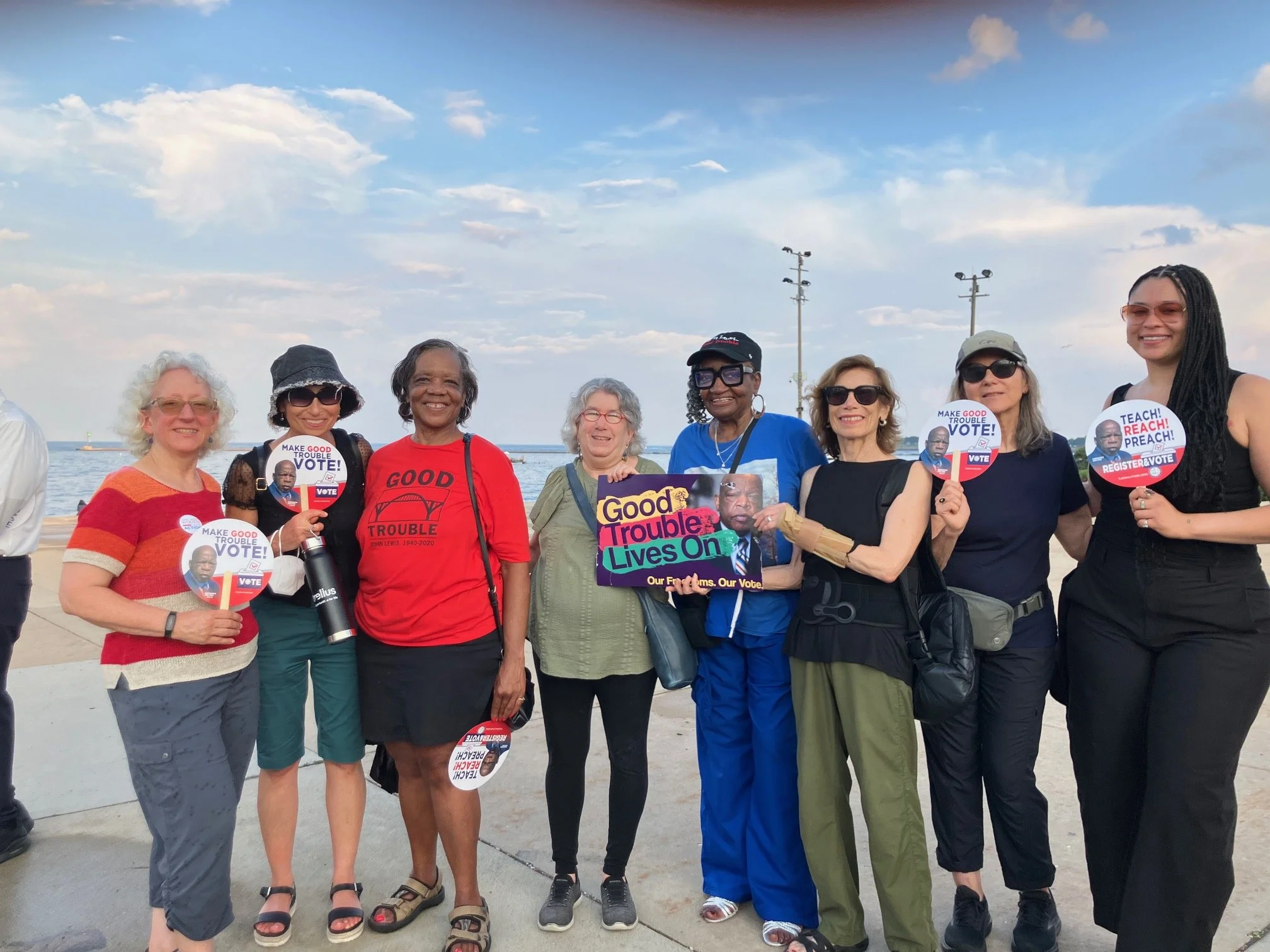 A group of diverse women standing together outdoors, smiling, holding political campaign signs supporting voting and political candidates, with a water body and a partly cloudy sky in the background.