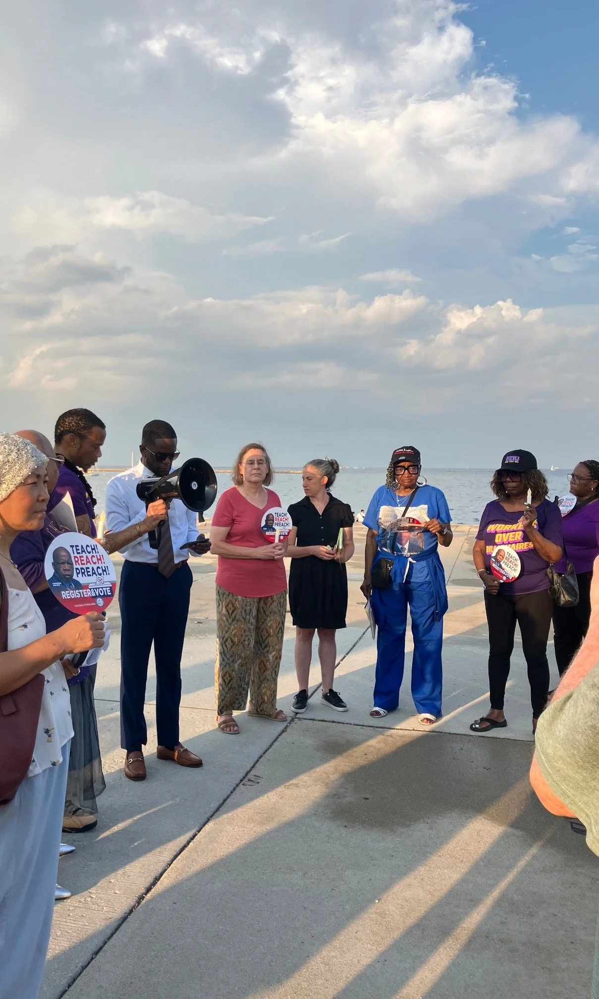 A diverse group of people gather outdoors near water under a partly cloudy sky, holding campaign signs and listening to a speaker with a megaphone.