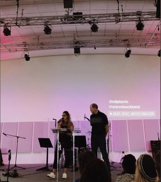 Two people standing on a stage with microphones, one woman and one man, in a large room with stage lighting and sound equipment, in Kent, United Kingdom.