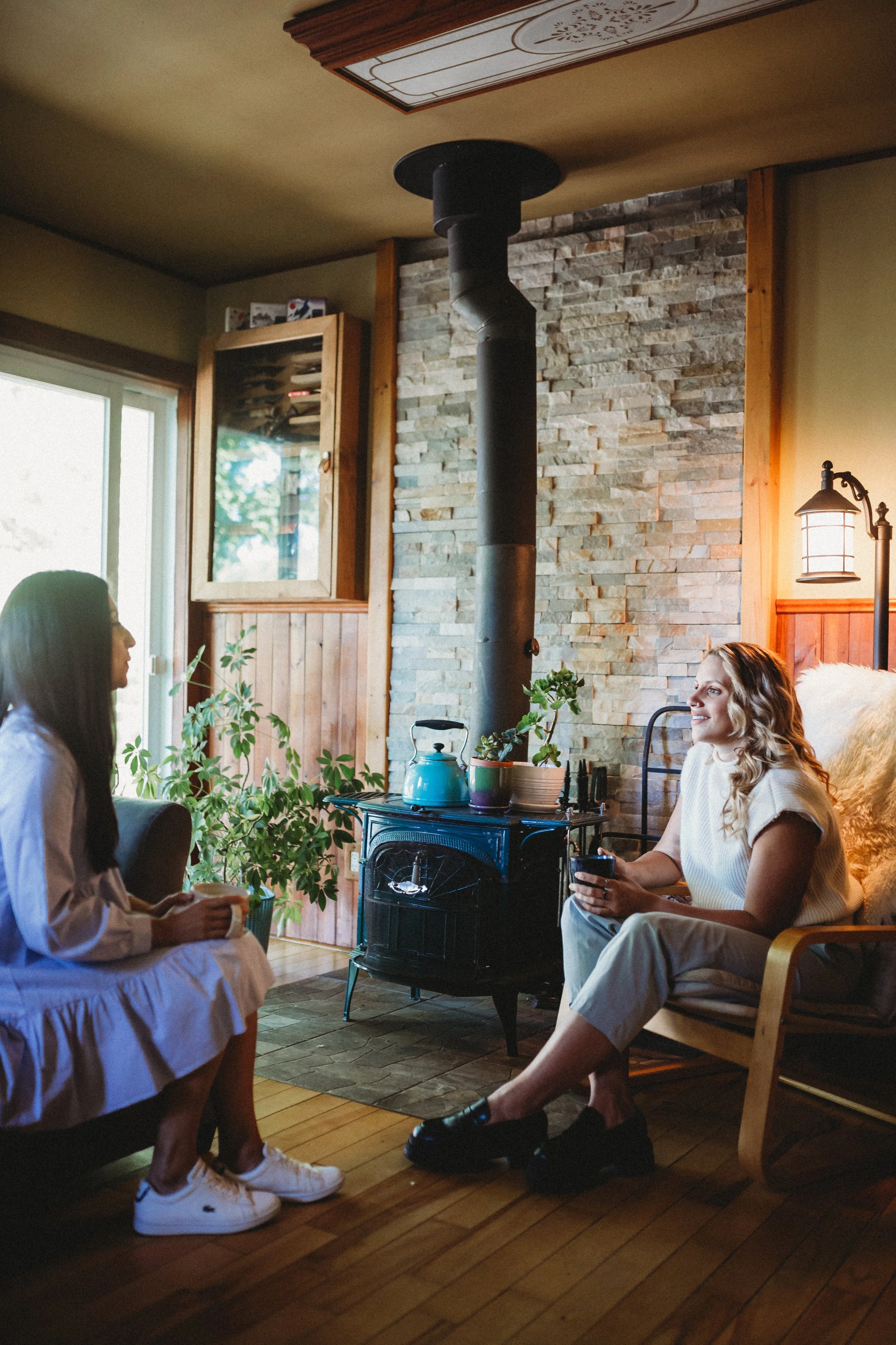 Two women sitting in a cozy, wood-paneled living room, talking near a wood stove with a visible chimney and a stone accent wall, during daytime.