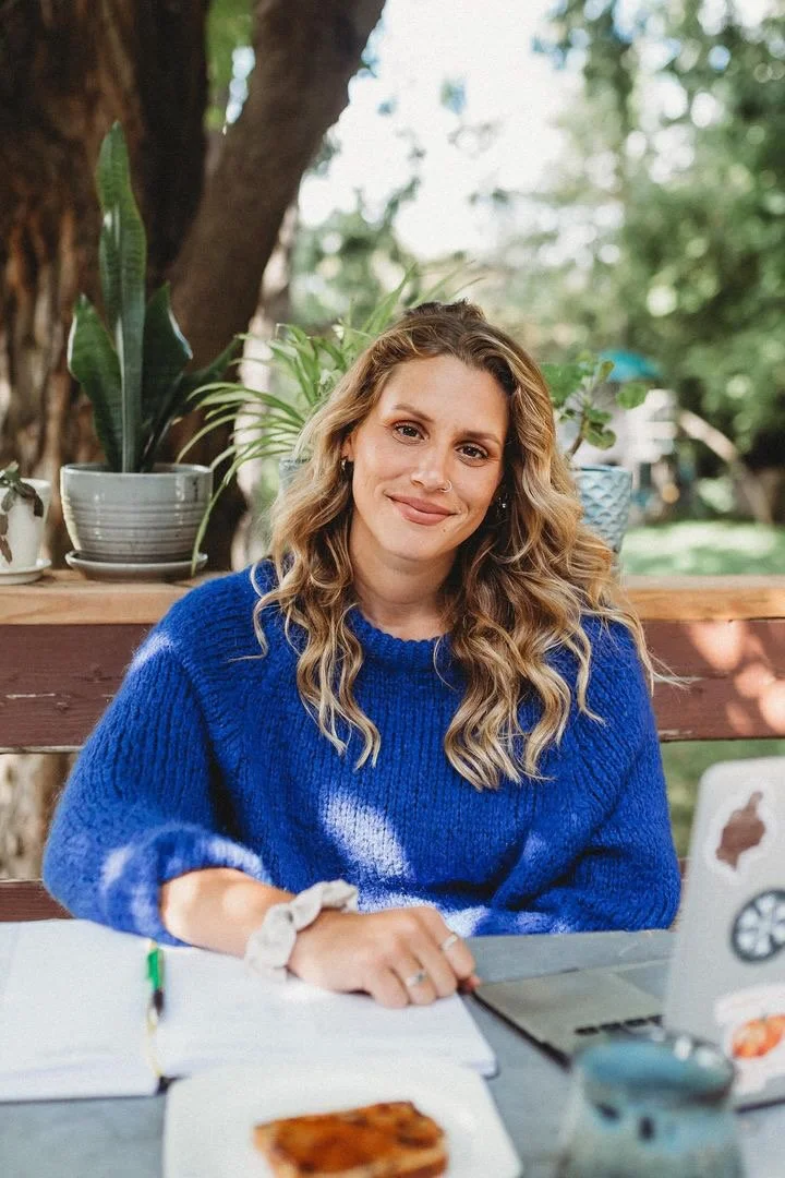 A woman with wavy blonde hair wearing a blue sweater, sitting outdoors at a table with a laptop, notebooks, and a plate with a cookie, smiling at the camera, with trees and plants in the background.