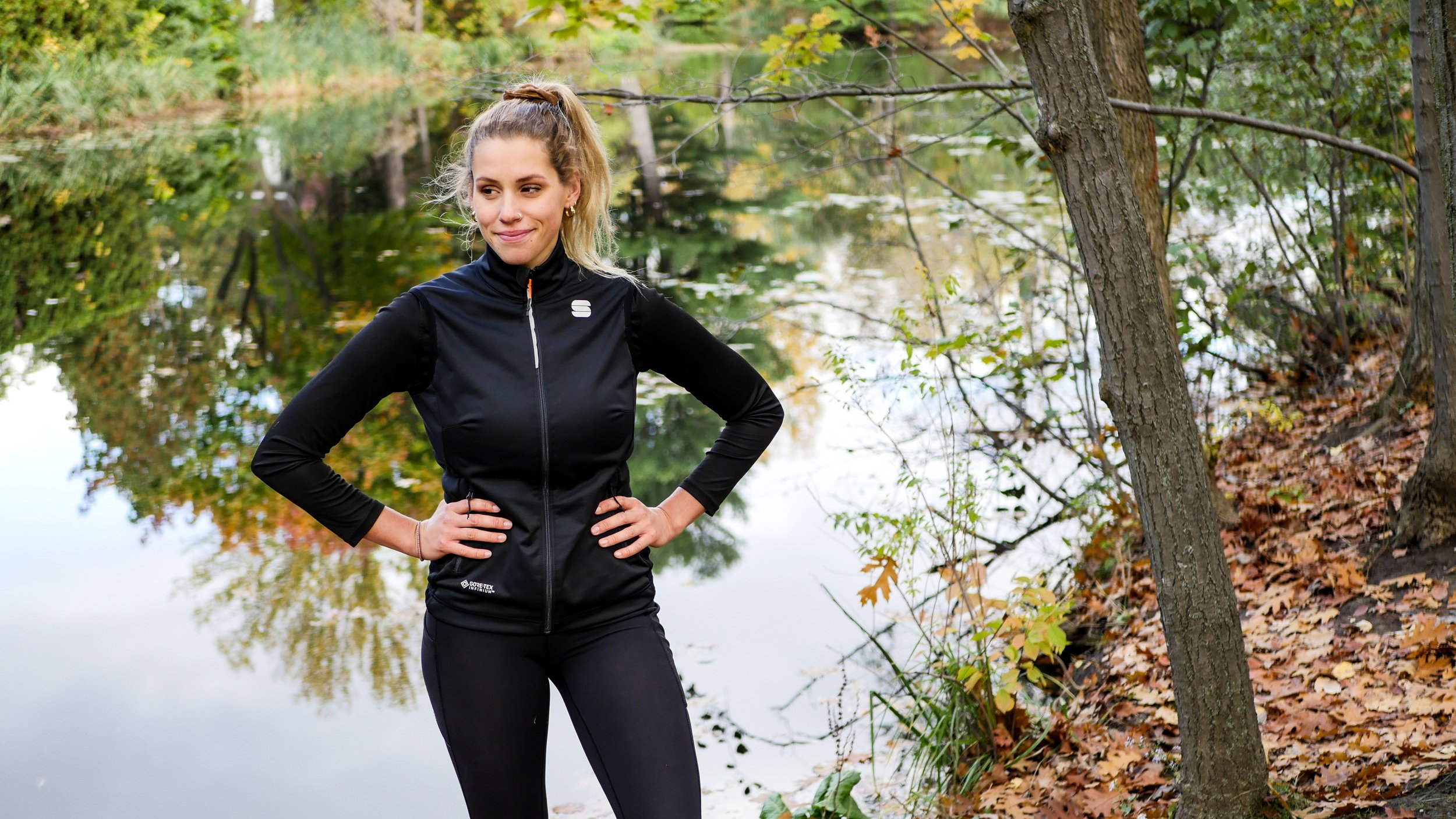 A woman in black athletic clothing standing outdoors near a lake in fall, with trees and fallen leaves around her.