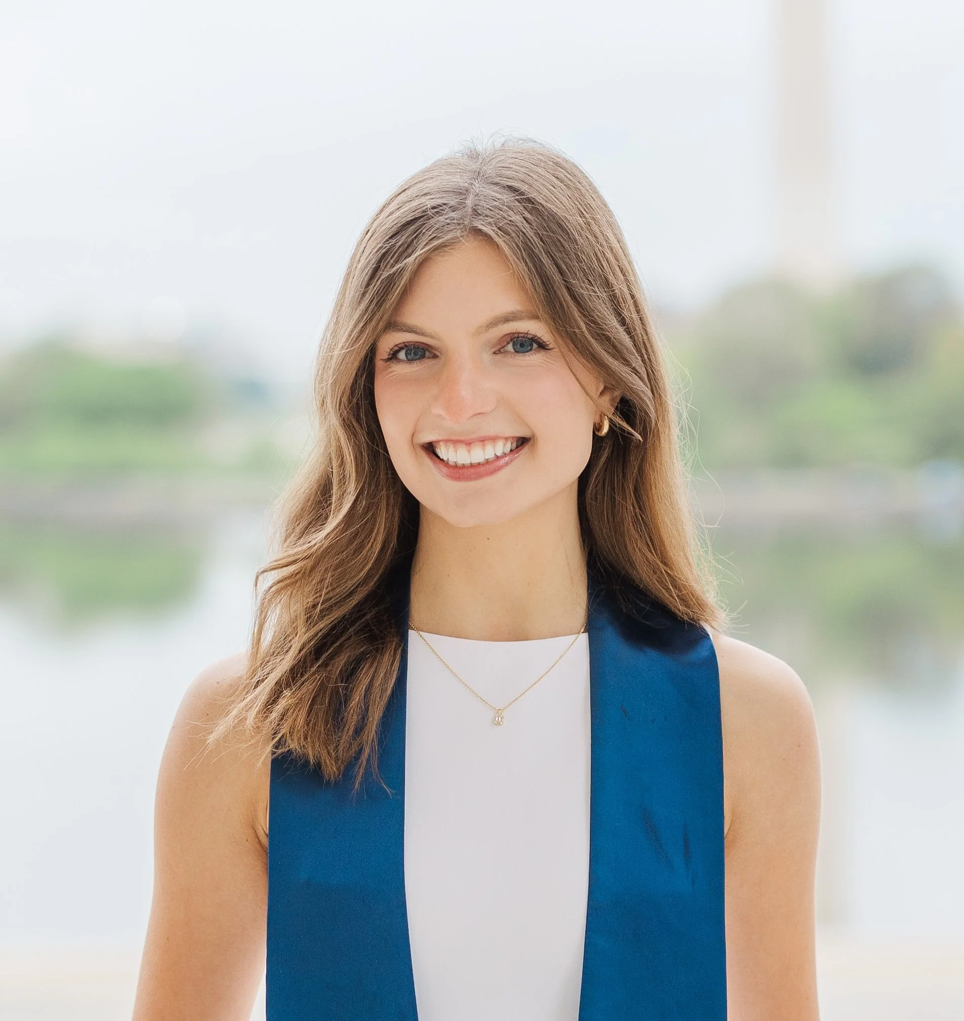 A young woman smiling with shoulder-length light brown hair, wearing a white top, a sleeveless blue vest, gold earrings, and a gold necklace, standing outdoors with a blurred background of water and greenery.