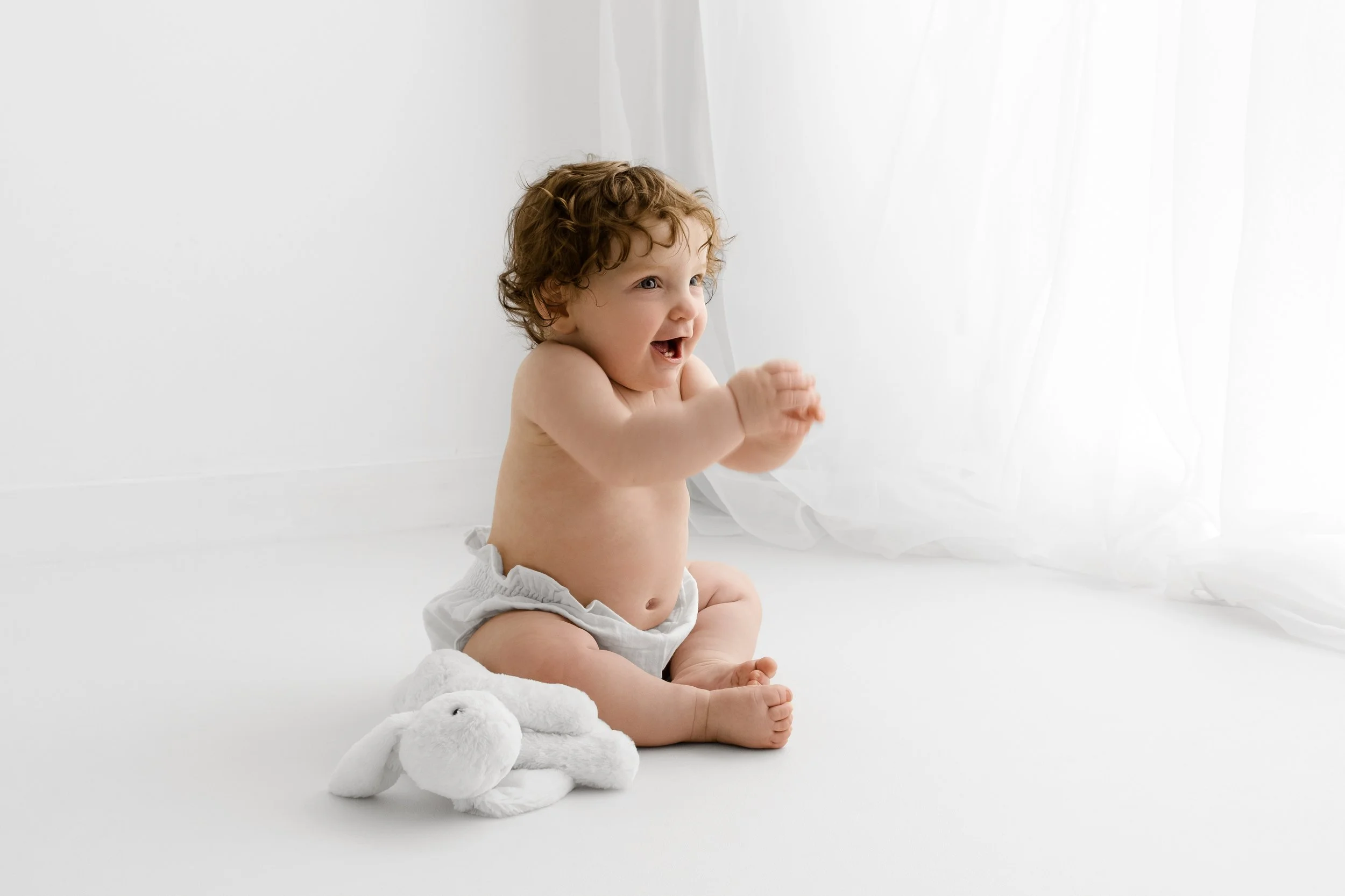 Curly haired 7 month old baby sitting and smiling in soft light at Dundee studio photoshoot