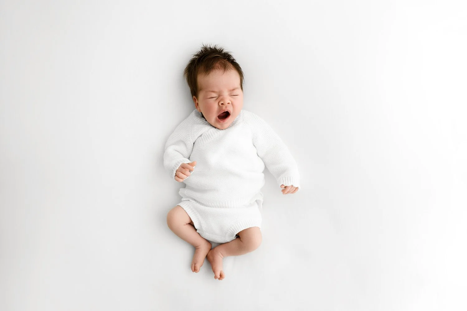 Newborn baby girl wearing a white knitted outfit, photographed on a soft neutral background to demonstrate timeless, minimalist newborn photography. baby is cute and yawning