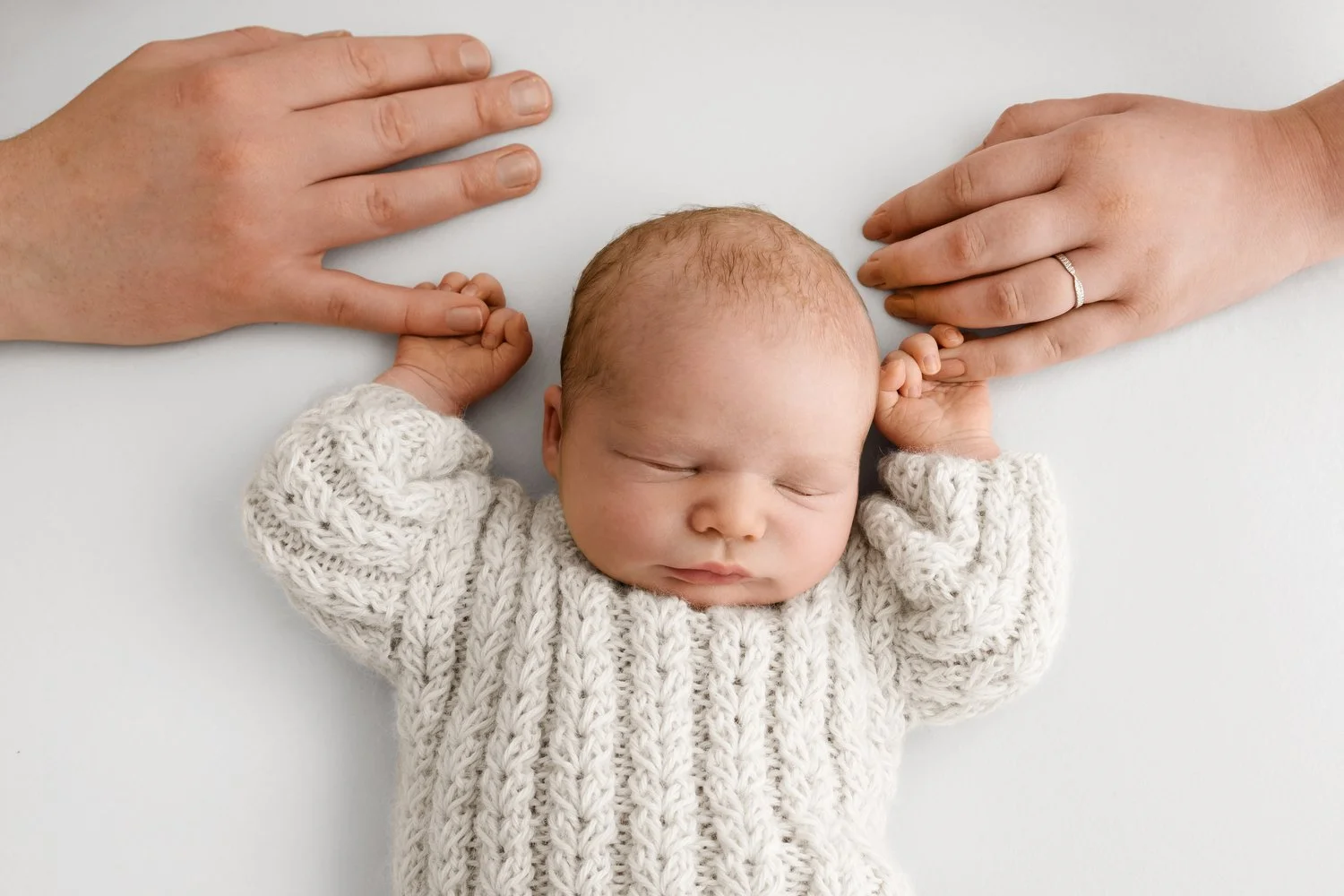 minimalist photo of newborn baby holding each parents finger. Baby is sleeping and is wearing a natural knitted sweater