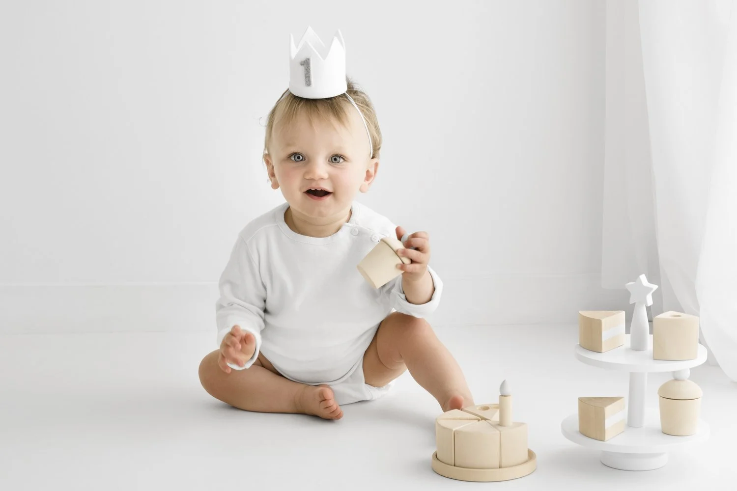 Baby wearing white outfit and felt crown playing with wooden birthday cake at first birthday photoshoot Mabel and Moose Dundee