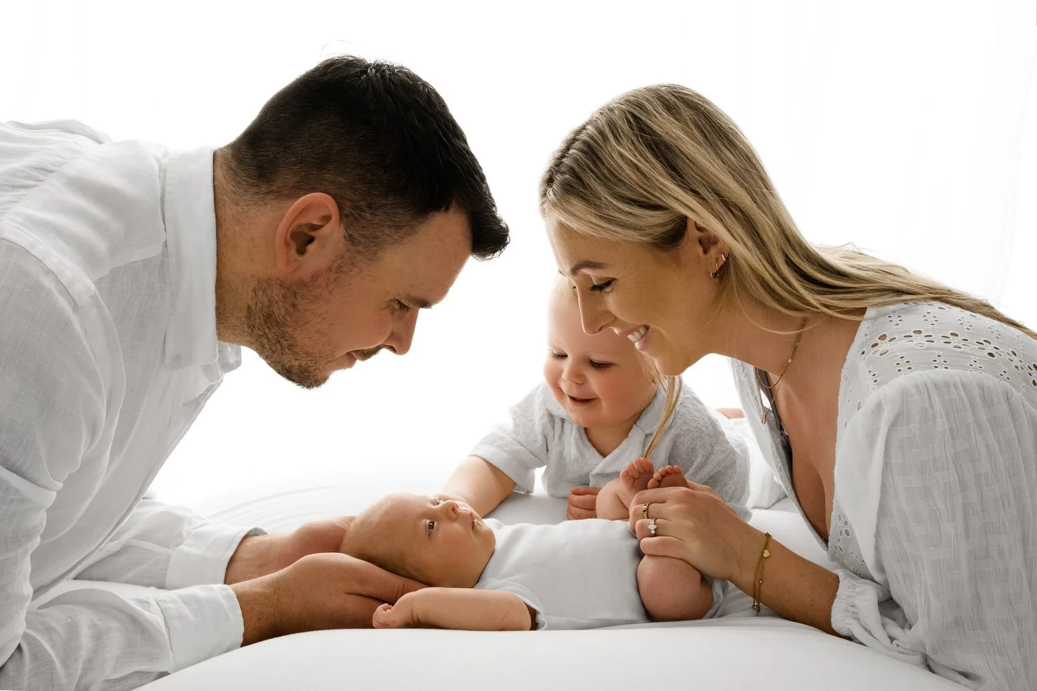 Parents and toddler looking down at their newborn during natural family photoshoot in Dundee studio, all wearing white