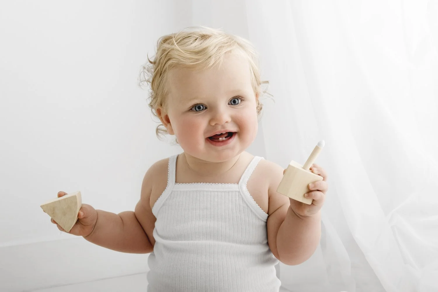 Smiling baby with blonde curly hair holding wooden birthday cake with one candle wearing white bodysuit at Mabel and Moose Dundee