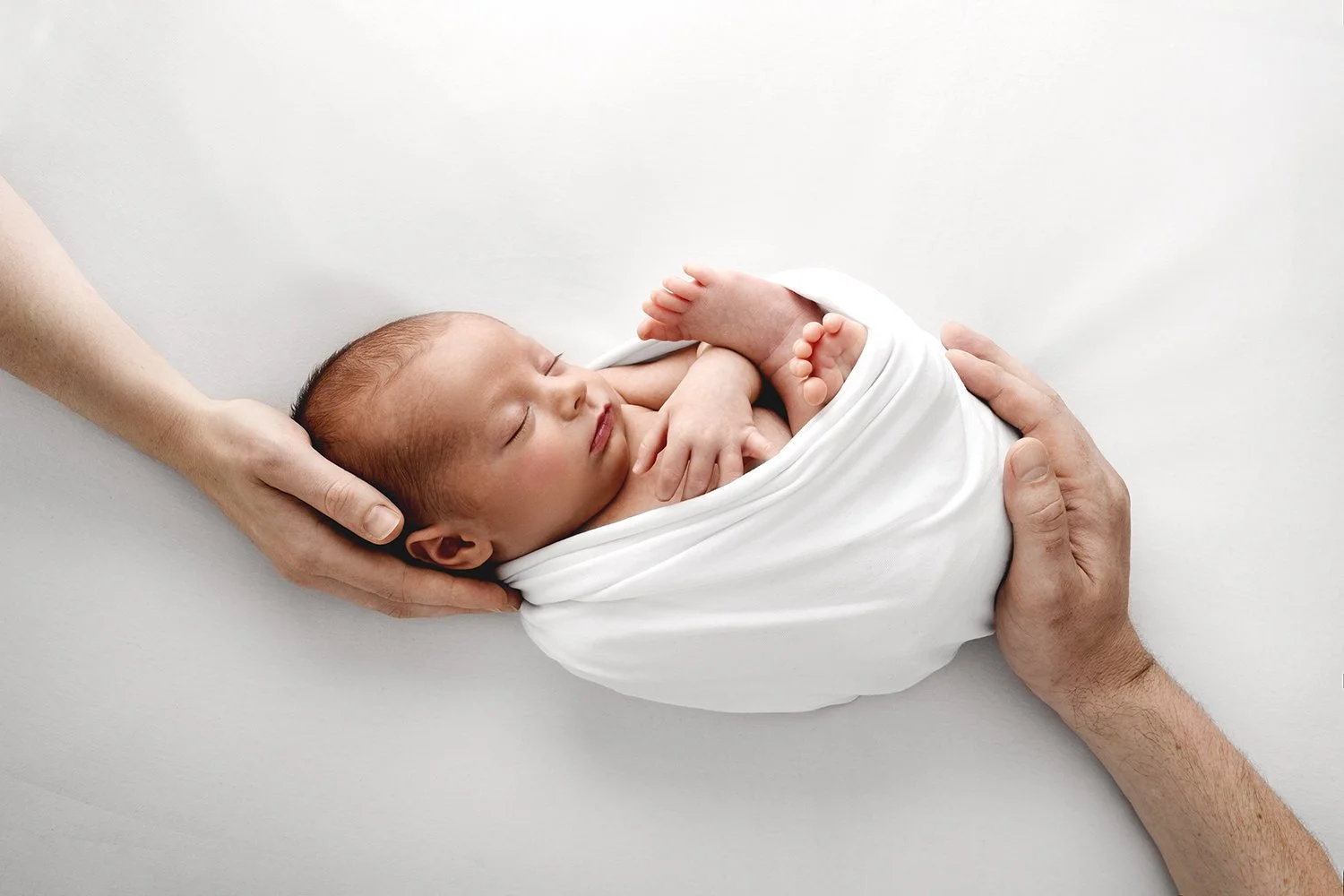 Newborn wrapped in white held gently in parents’ hands at Dundee photography studio