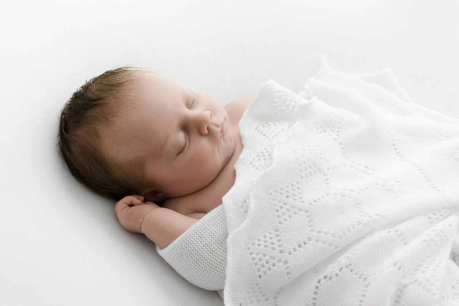 unposed newborn baby wrapped in a white knitted shawl. baby looks peaceful and is sleeping