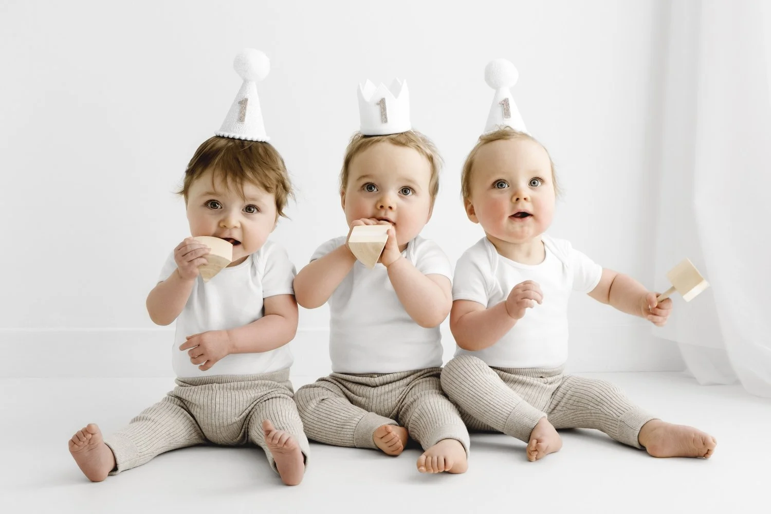 First birthday triplets wearing one hats and holding wooden cake slices at Dundee studio photoshoot