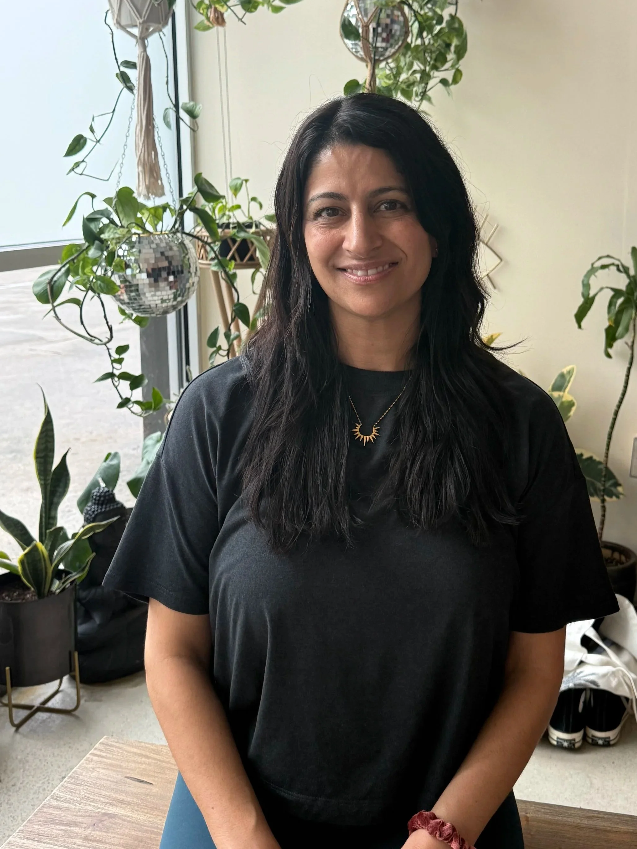 A woman with dark hair, wearing gold earrings and a dark sleeveless top, smiling and holding her hands in a prayer position outdoors.