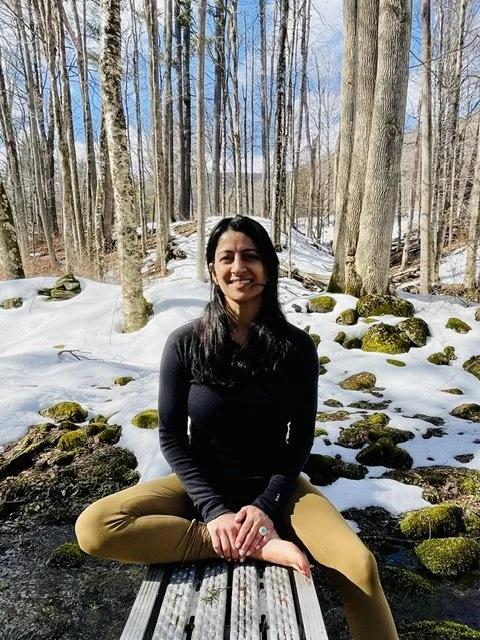 A woman with dark hair, wearing gold earrings and a dark sleeveless top, smiling and holding her hands in a prayer position outdoors.