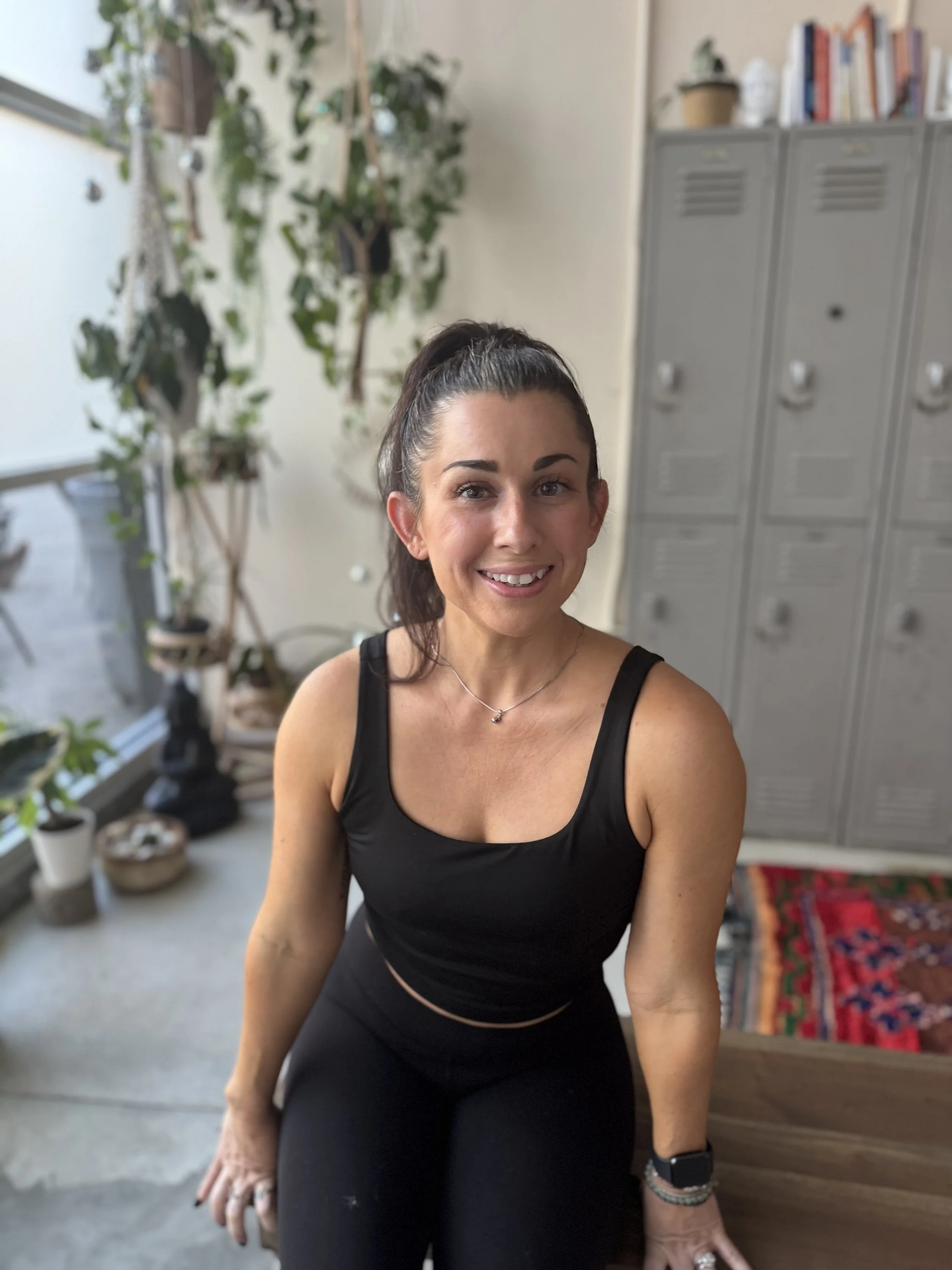 A smiling woman with shoulder-length wavy brown hair, wearing a black t-shirt and a delicate necklace, sitting in a room with a mirror and indoor plants in the background.