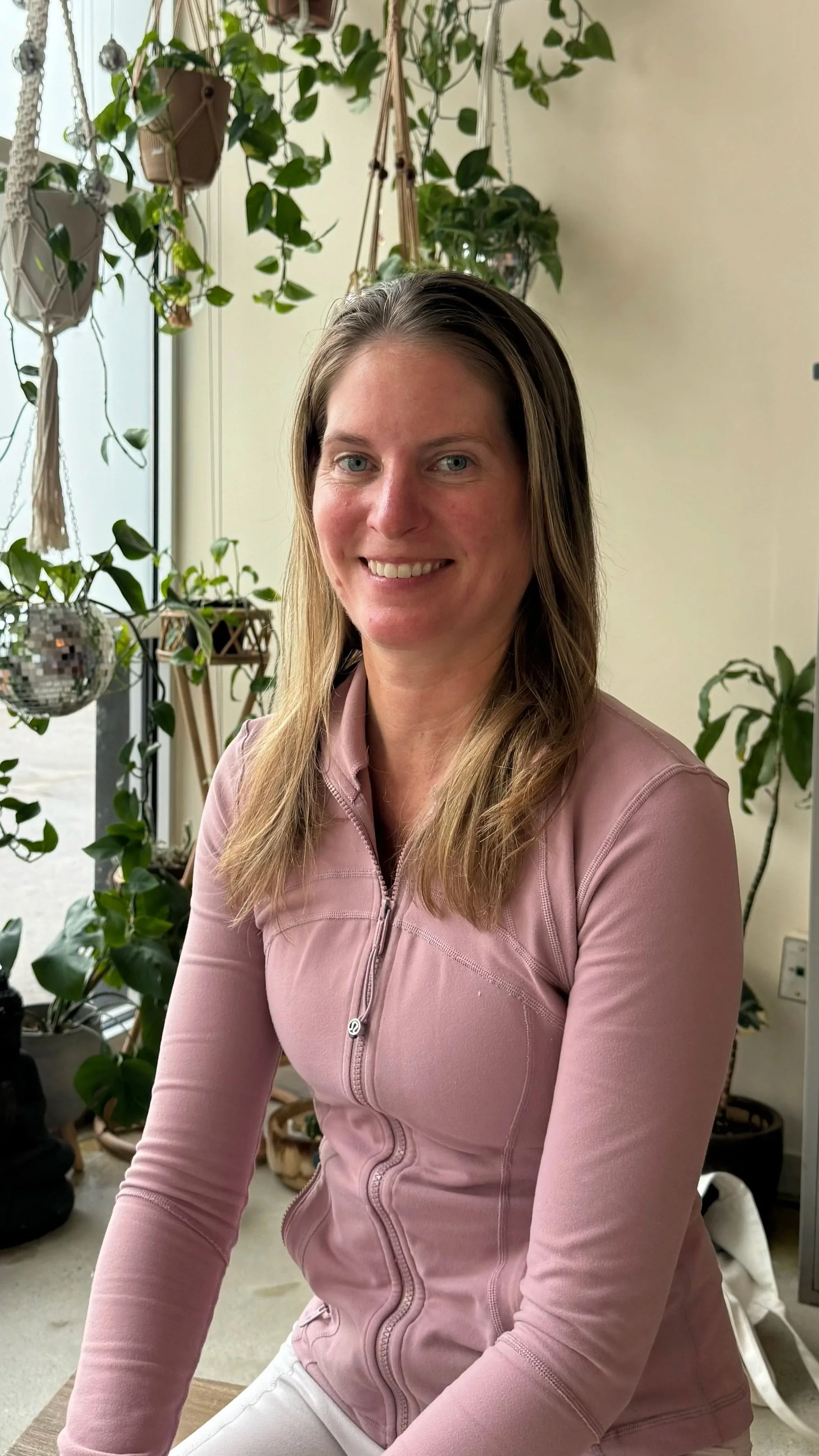 A woman with long hair smiling and practicing yoga or dance with her arms raised against a brick wall inside a room with large windows.