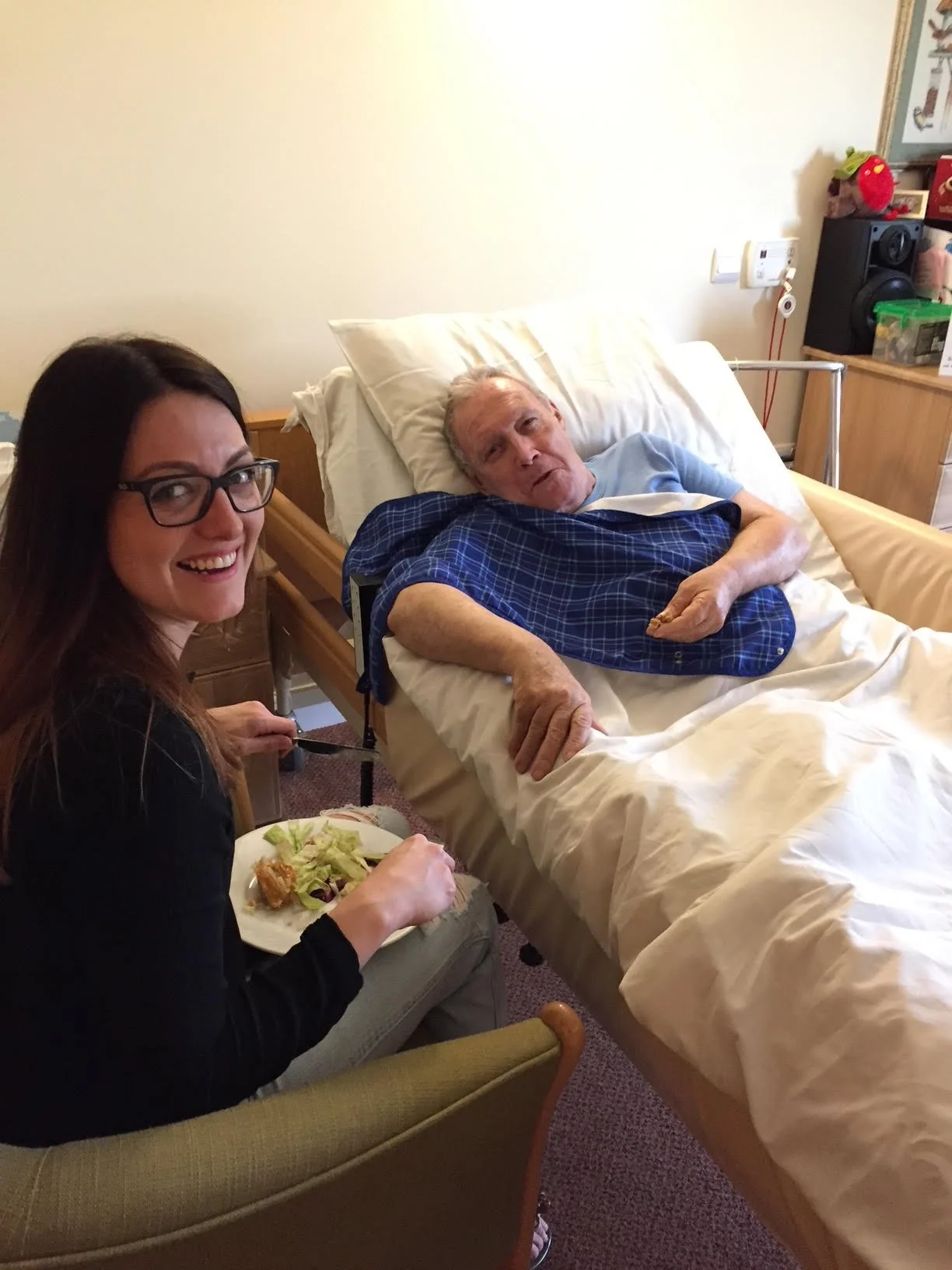 A woman with glasses smiling while sitting in a chair and holding a plate of food next to an elderly man in a hospital bed with white sheets, wearing a blue hospital gown, in a room with medical equipment and a stuffed red animal on a shelf.