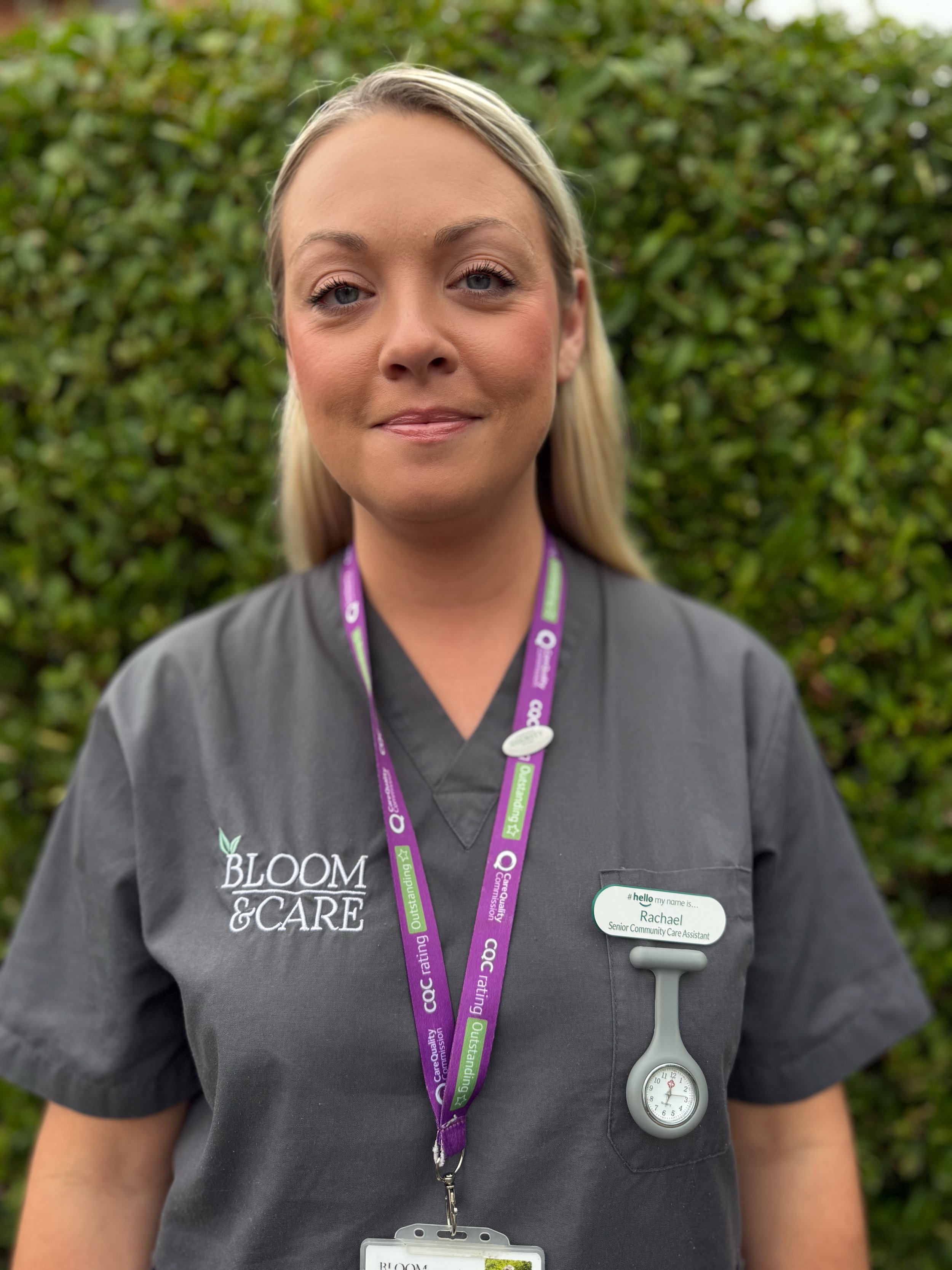 A woman with light blonde hair in a bun, wearing a light blue shirt and a name tag, standing outdoors with green foliage in the background.