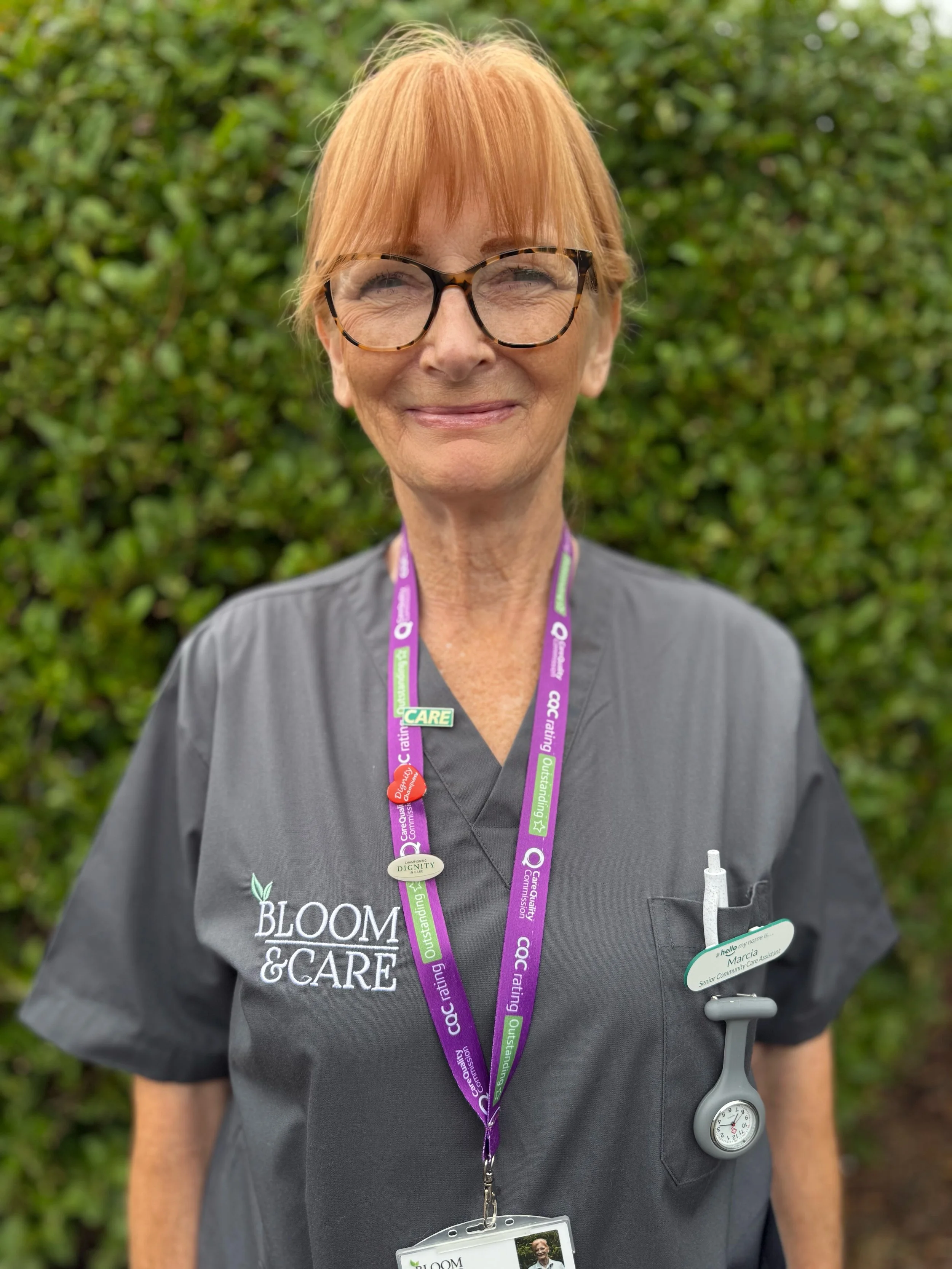 A woman with short, light auburn hair and fair skin, standing outdoors in front of green foliage, wearing a white striped uniform shirt with a name tag and a purple lanyard with green accents.