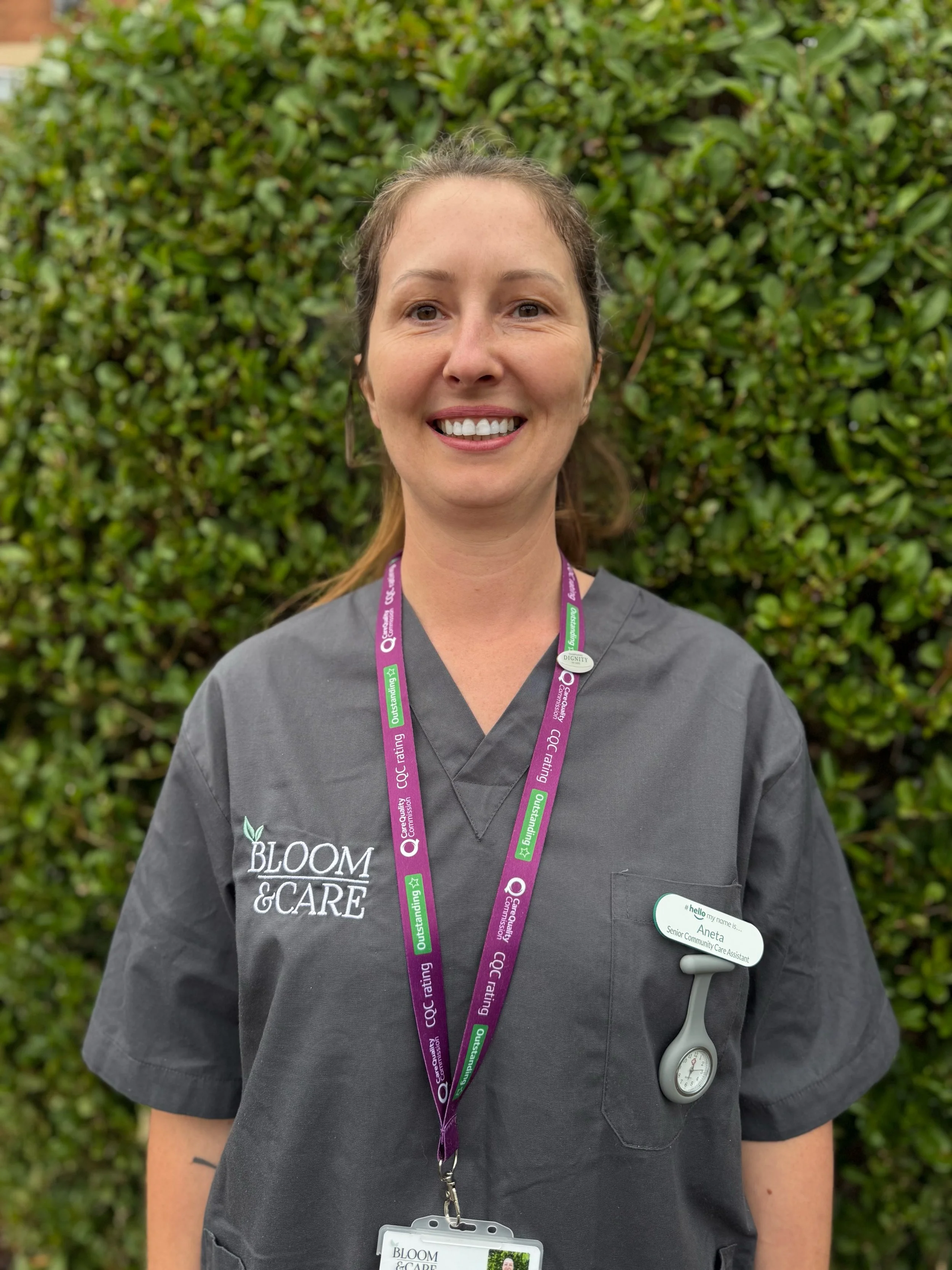 A woman with short brown hair wearing a light blue uniform with a name tag that reads 'Aneta, Community Care Assistant,' standing outdoors with green bushes in the background.
