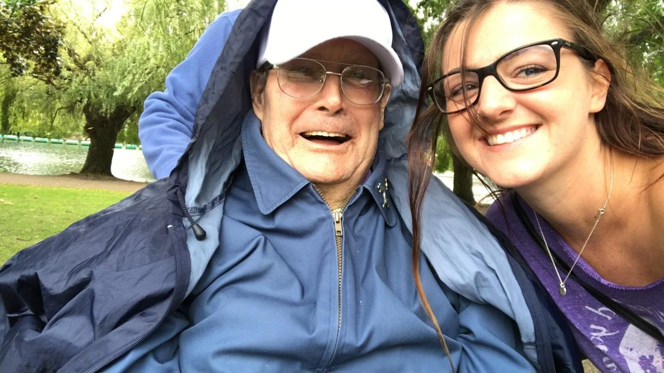 A smiling young woman with glasses and a necklace taking a selfie with an elderly man with glasses, wearing a blue jacket and cap, outdoors near a lake and trees.