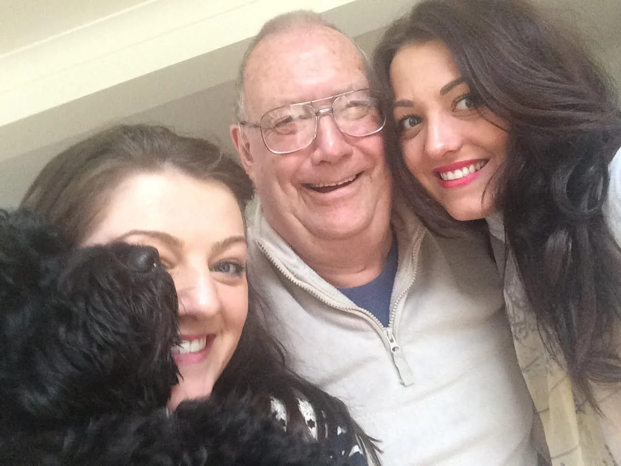 A group selfie of three women and a dog. The women have dark hair, and the man in the middle has glasses and light skin. They are smiling and close together.
