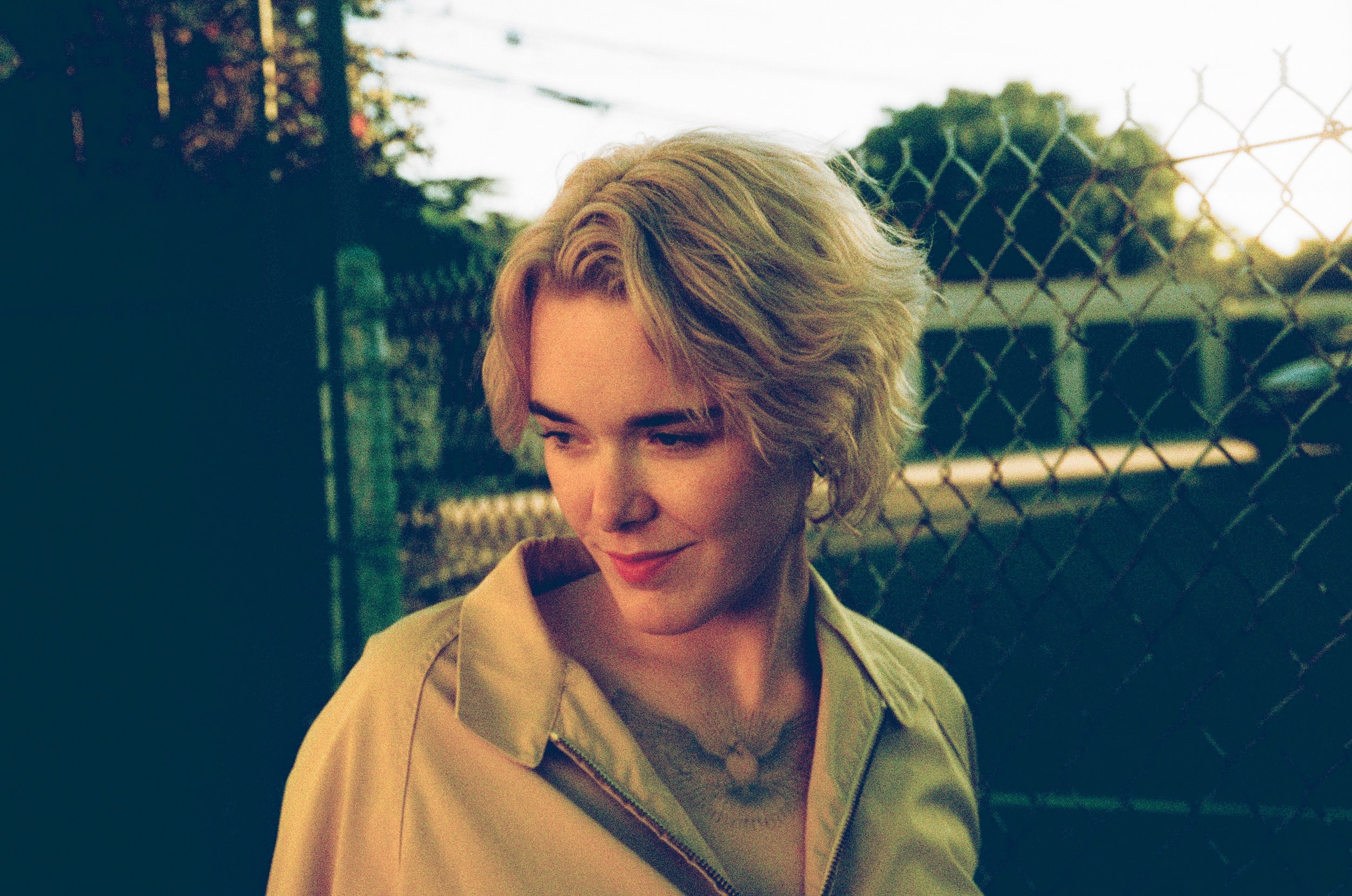 A woman with short, curly blonde hair, wearing a light-colored jacket, standing outdoors near a chain-link fence during sunset.