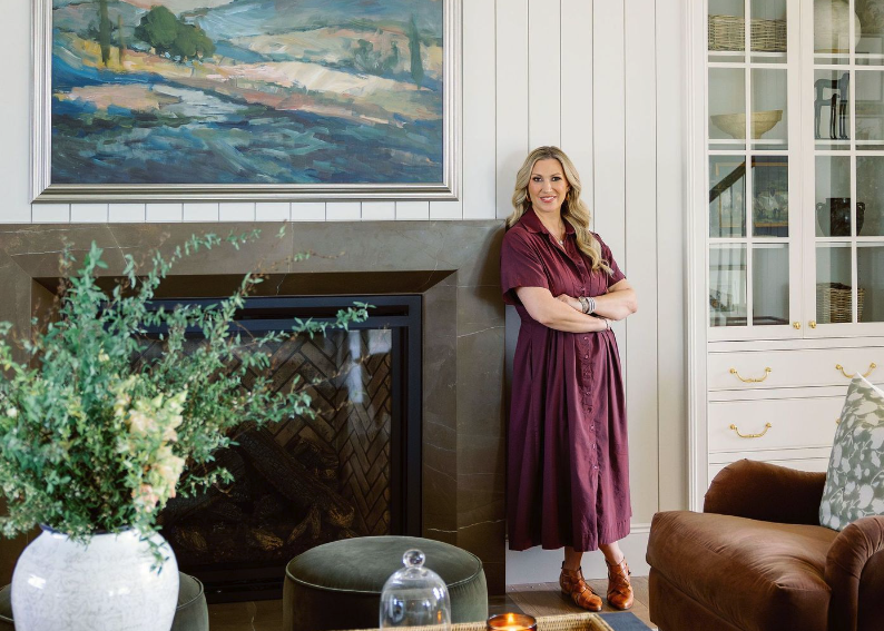 A woman in a maroon dress standing next to a fireplace in a living room, smiling with arms crossed. There is a painting above the fireplace, a large plant in a white pot, a brown armchair with a patterned pillow, and a glass cabinet with decor inside.