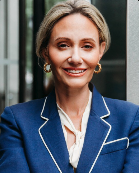 A woman with short blonde hair, wearing a blue blazer with white piping and gold earrings, smiling in an outdoor setting with glass buildings in the background.