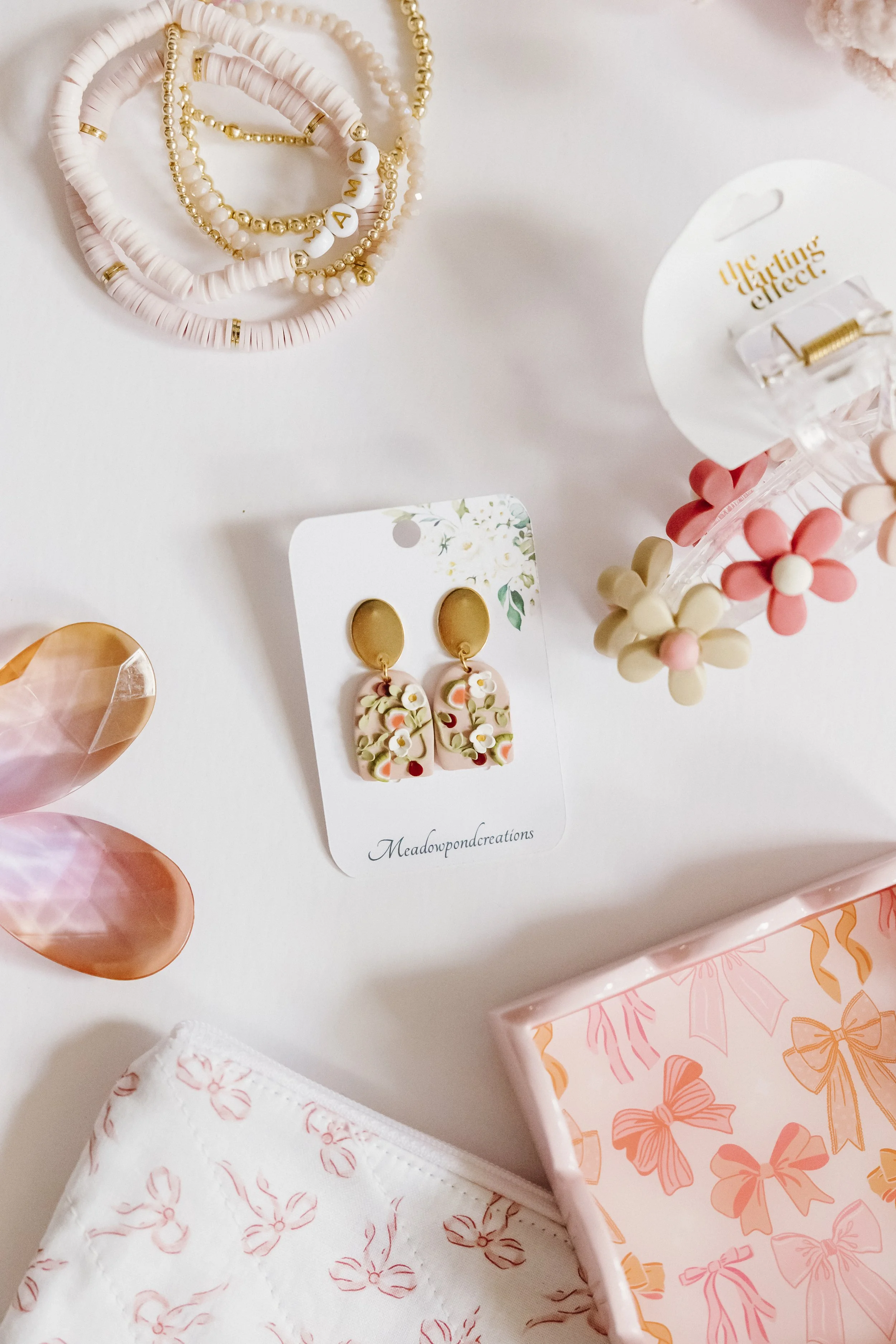 Pink jewelry, floral earrings on a card, pink hair clips with flower shapes, pink gift box with bows pattern, pink and white pouch, and pink shell hair accessory on a white surface.