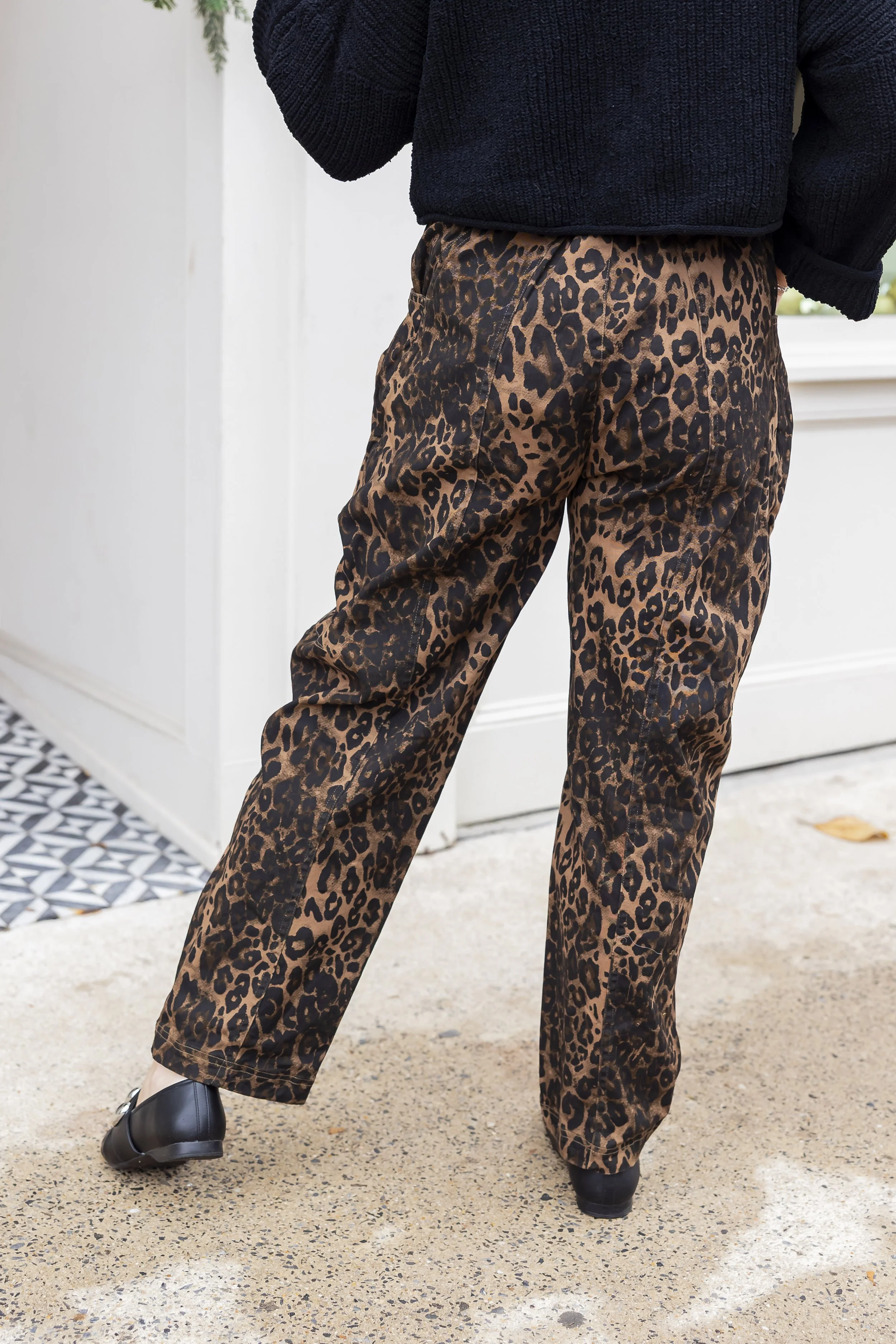 Person wearing leopard print pants and a black sweater, standing outside near a white wall and patterned tile.