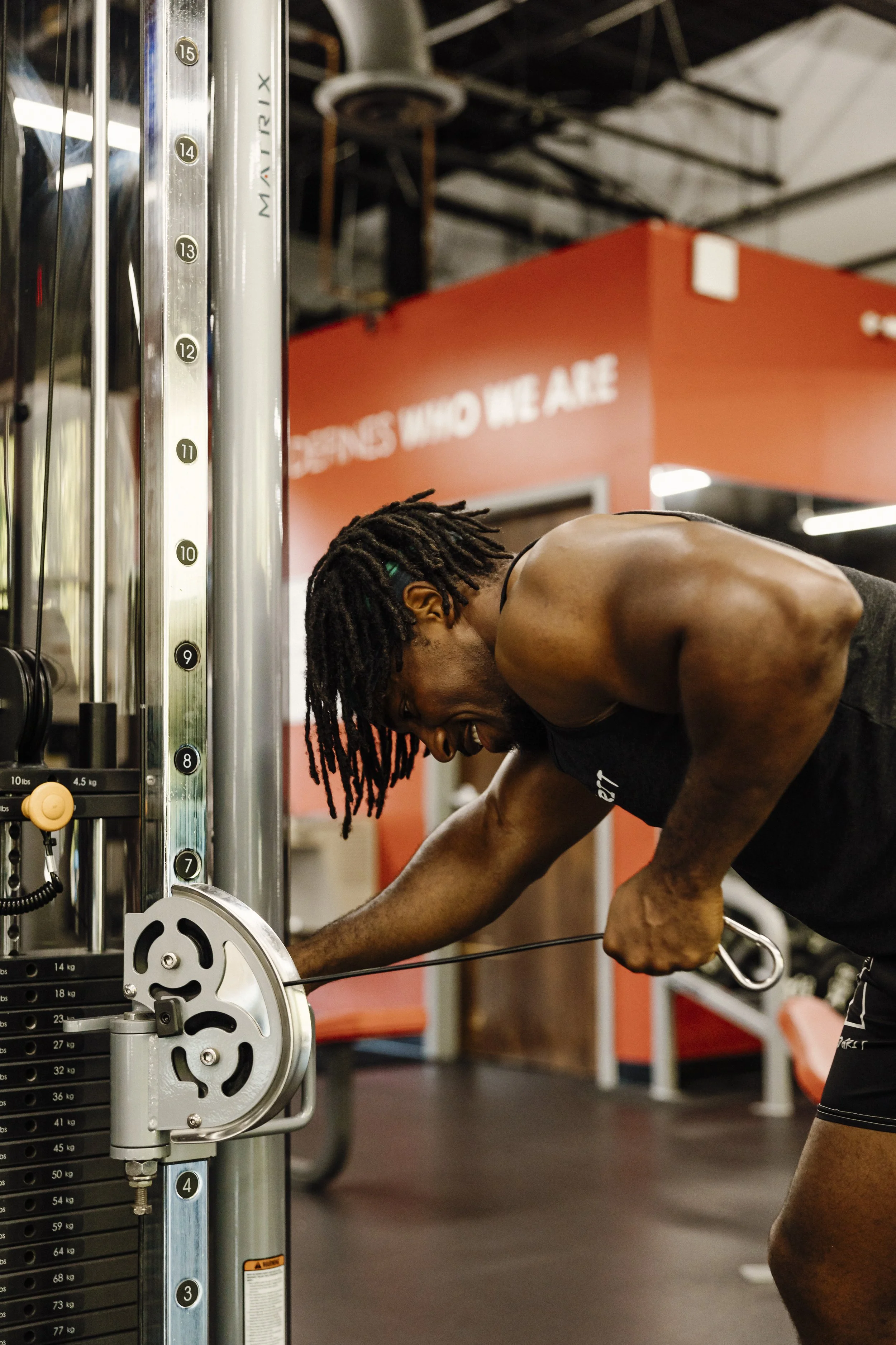 A shirtless man with dreadlocks working out on a machine in a gym, smiling and leaning forward.