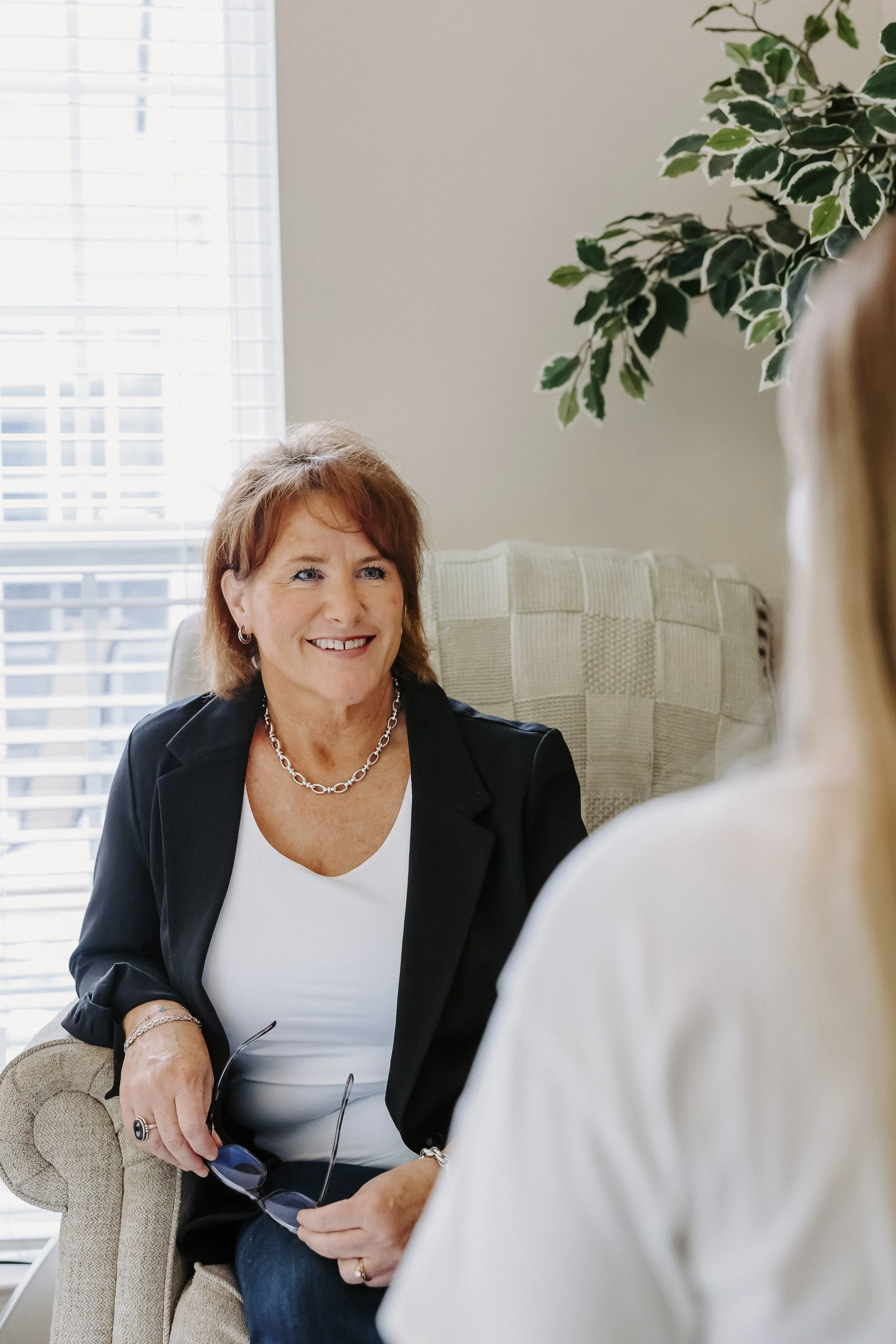 A woman with short red hair wearing a black blazer and white top, smiling while sitting on an armchair and holding glasses, engaged in conversation with another woman whose back is to the camera.