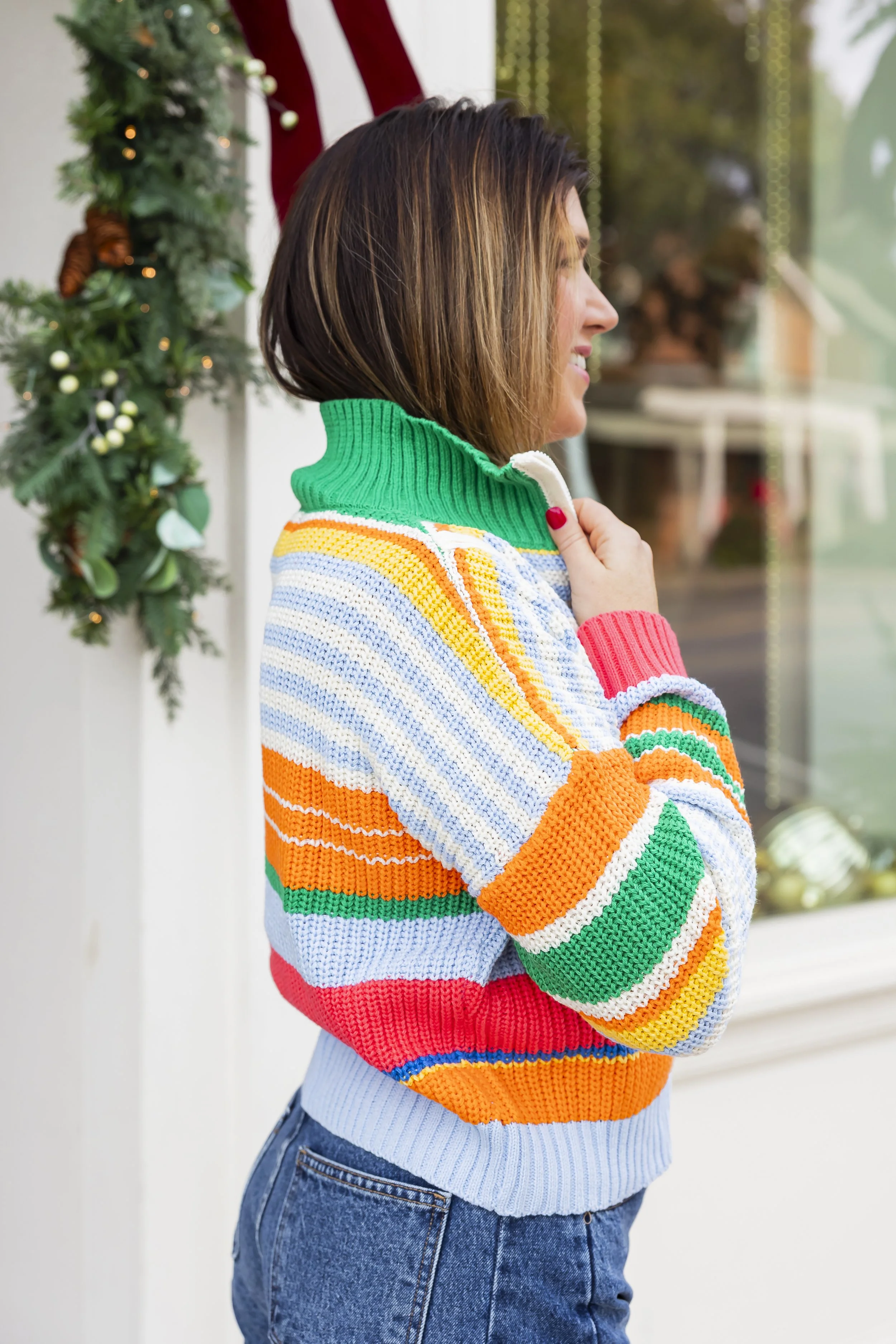 A woman stands outside near a decorated holiday window display, wearing a colorful, striped sweater with a green turtleneck, and has short brown hair.