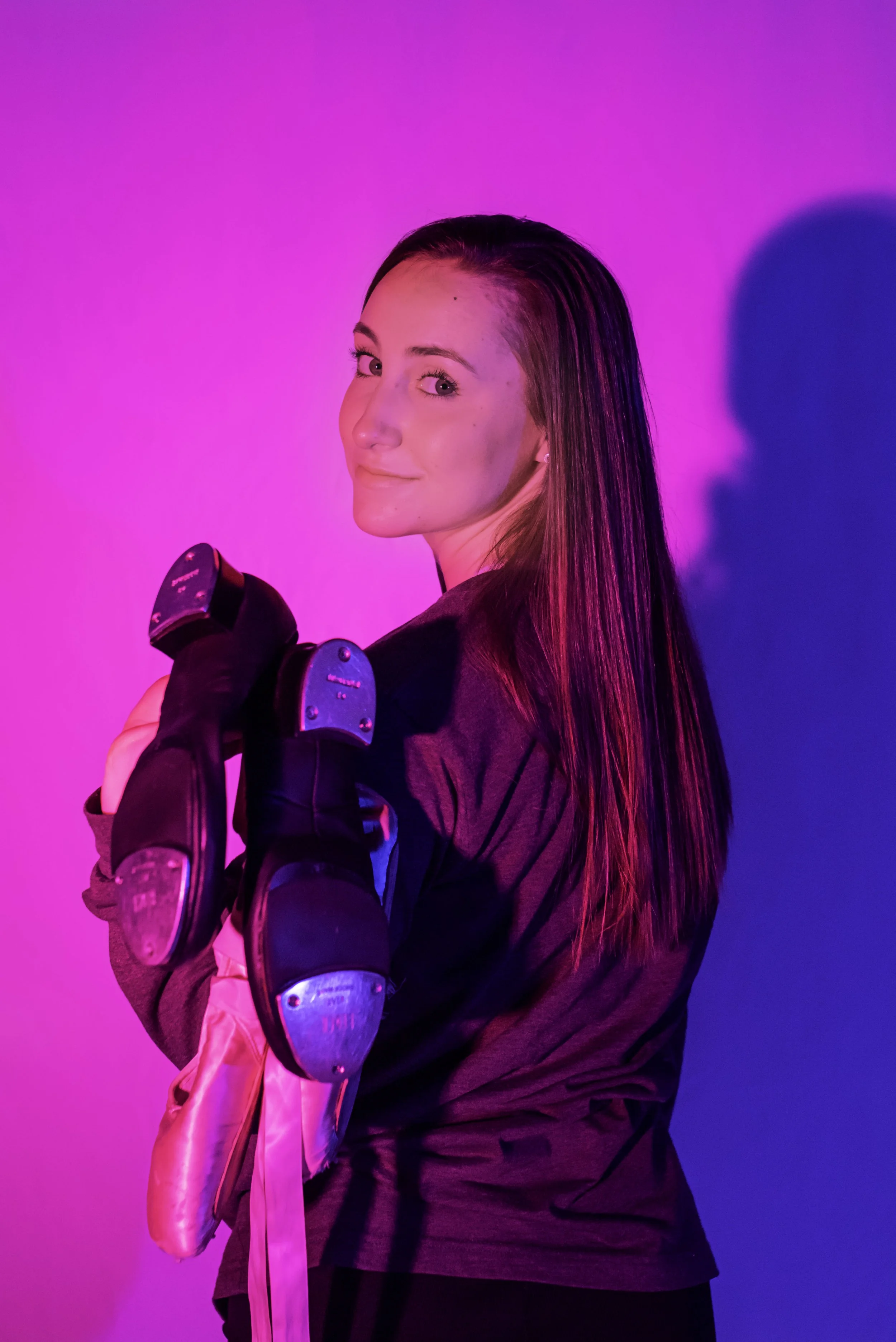 A woman with long straight hair holding a pair of rollerblades, standing against a vibrant pink and purple background with a shadow cast on the wall.