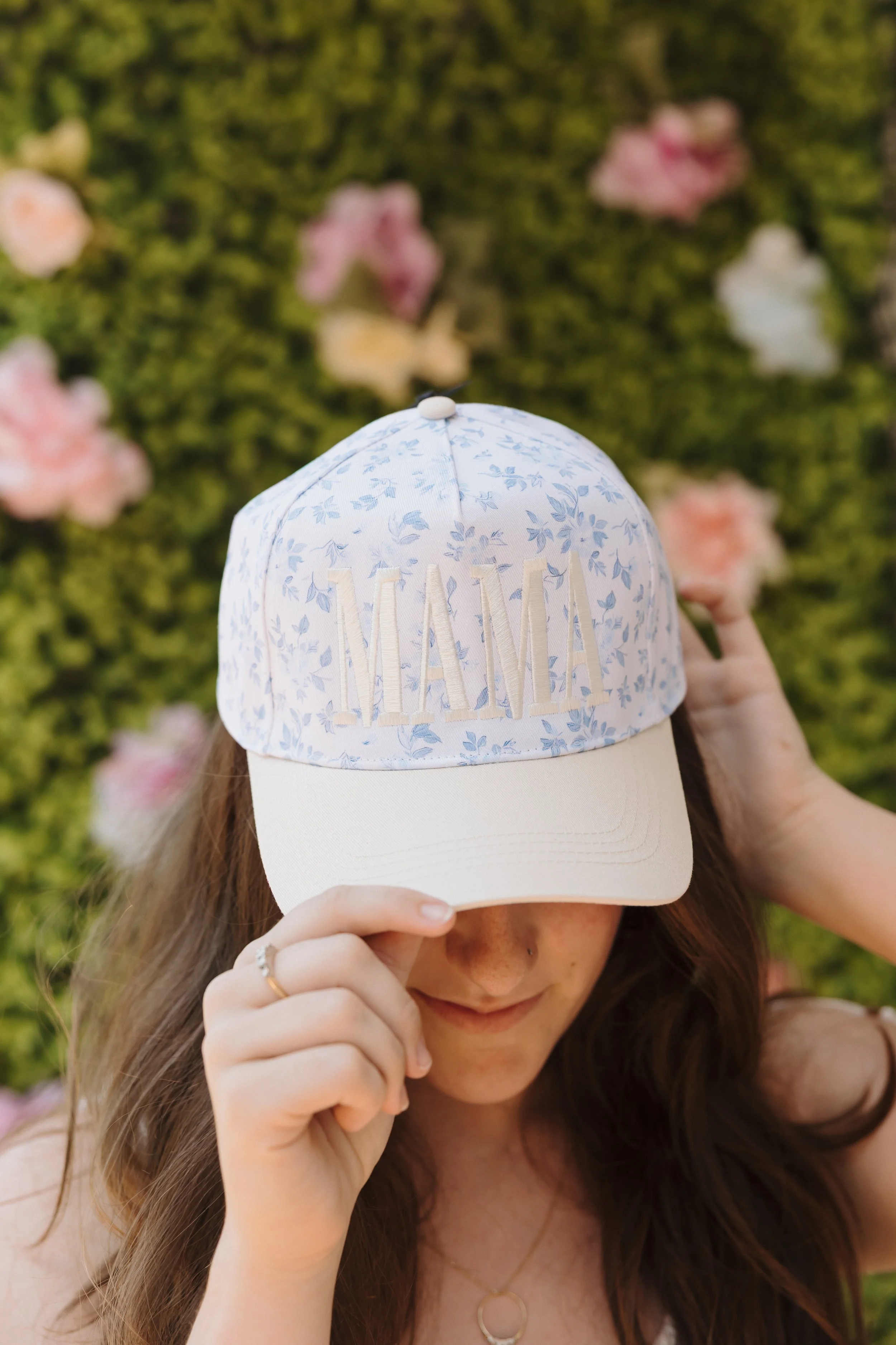 A woman wearing a light-colored cap with floral patterns and the word 'MIAMI' embroidered on it. She is adjusting the cap, with her face partially obscured, standing in front of a green, flower-covered background.