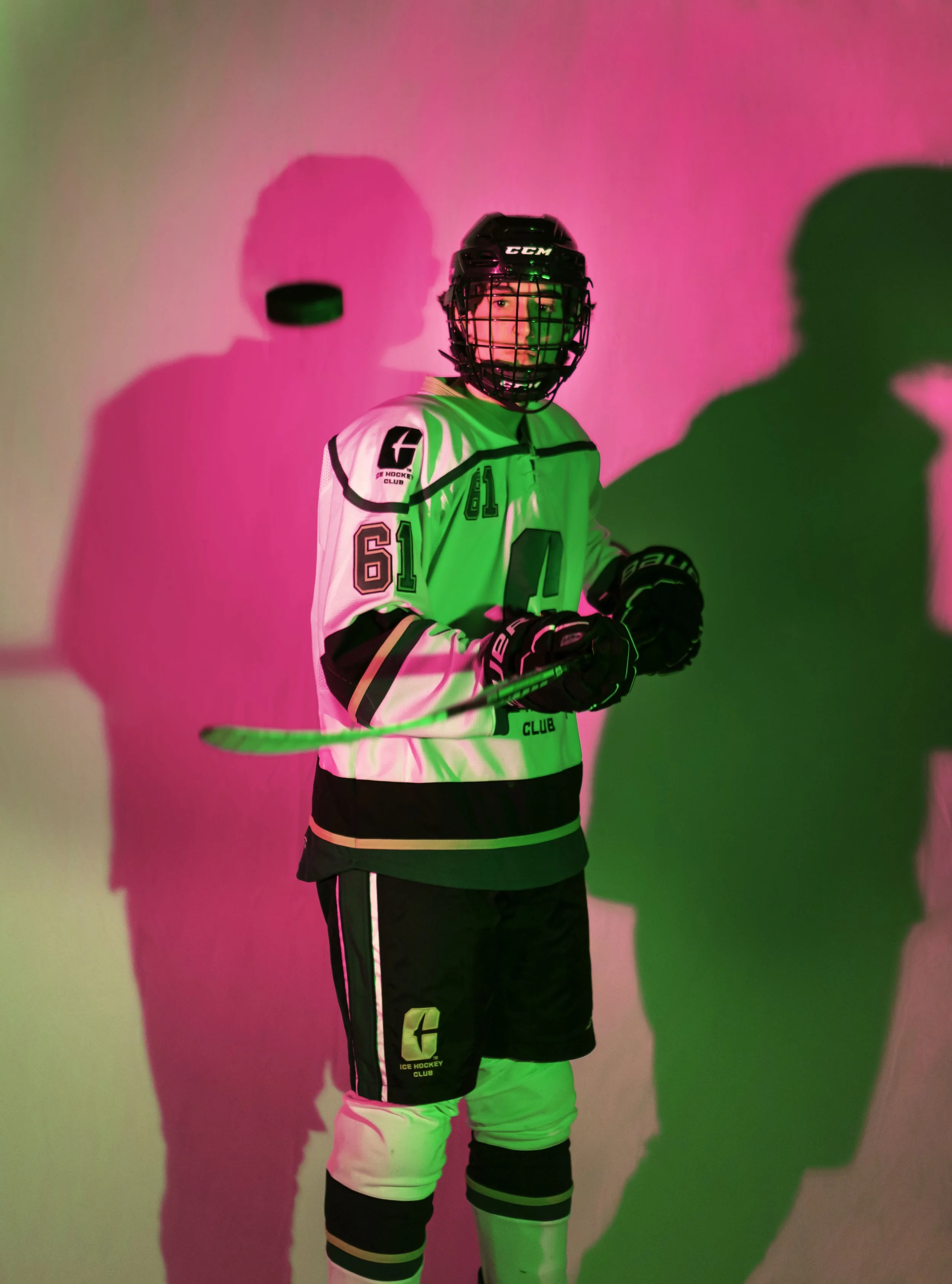 An ice hockey player in full gear holding a hockey stick, standing against a pink and green background with shadows cast by the lighting.