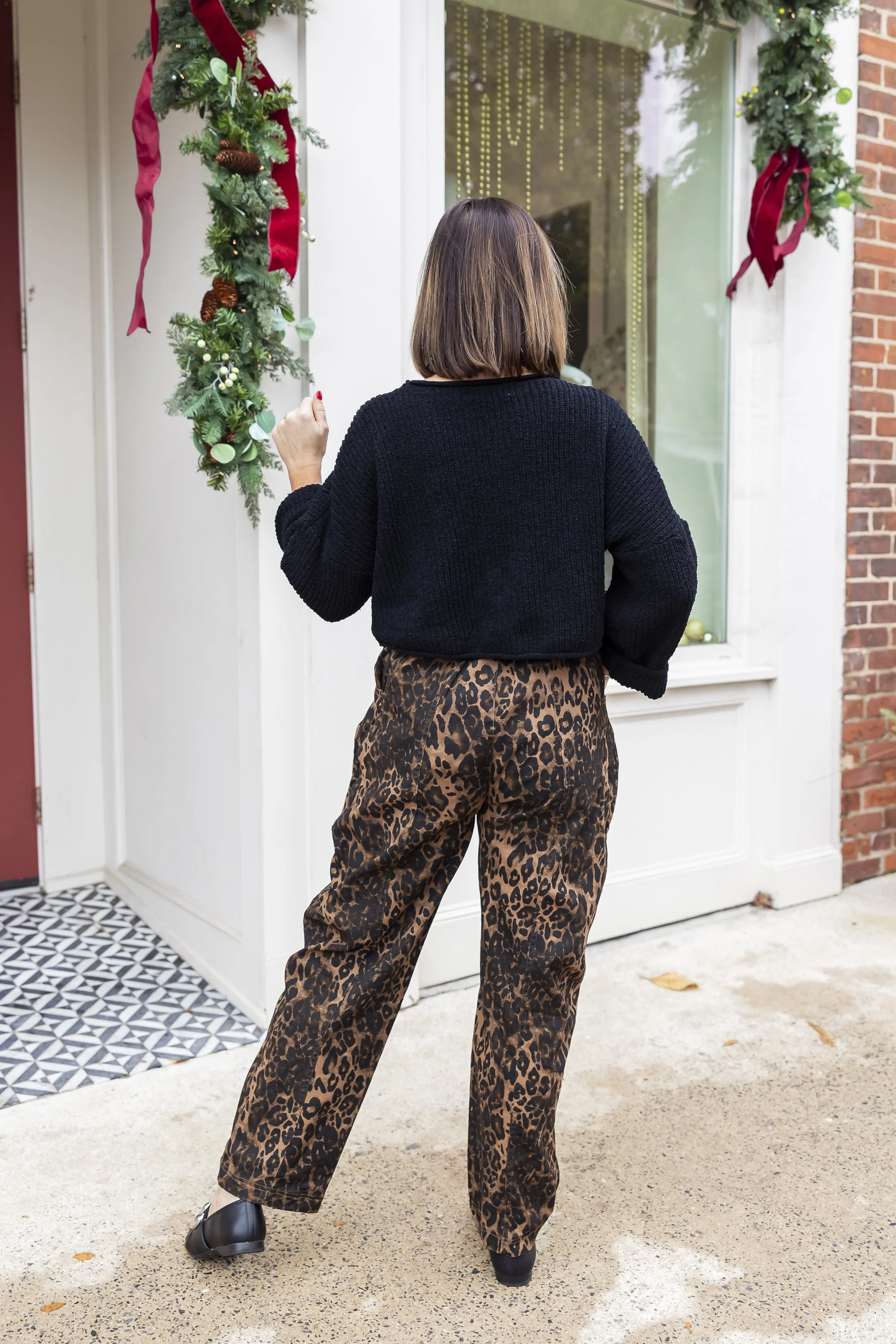A woman standing outside a house, facing a decorated door for Christmas with greenery and red ribbons.