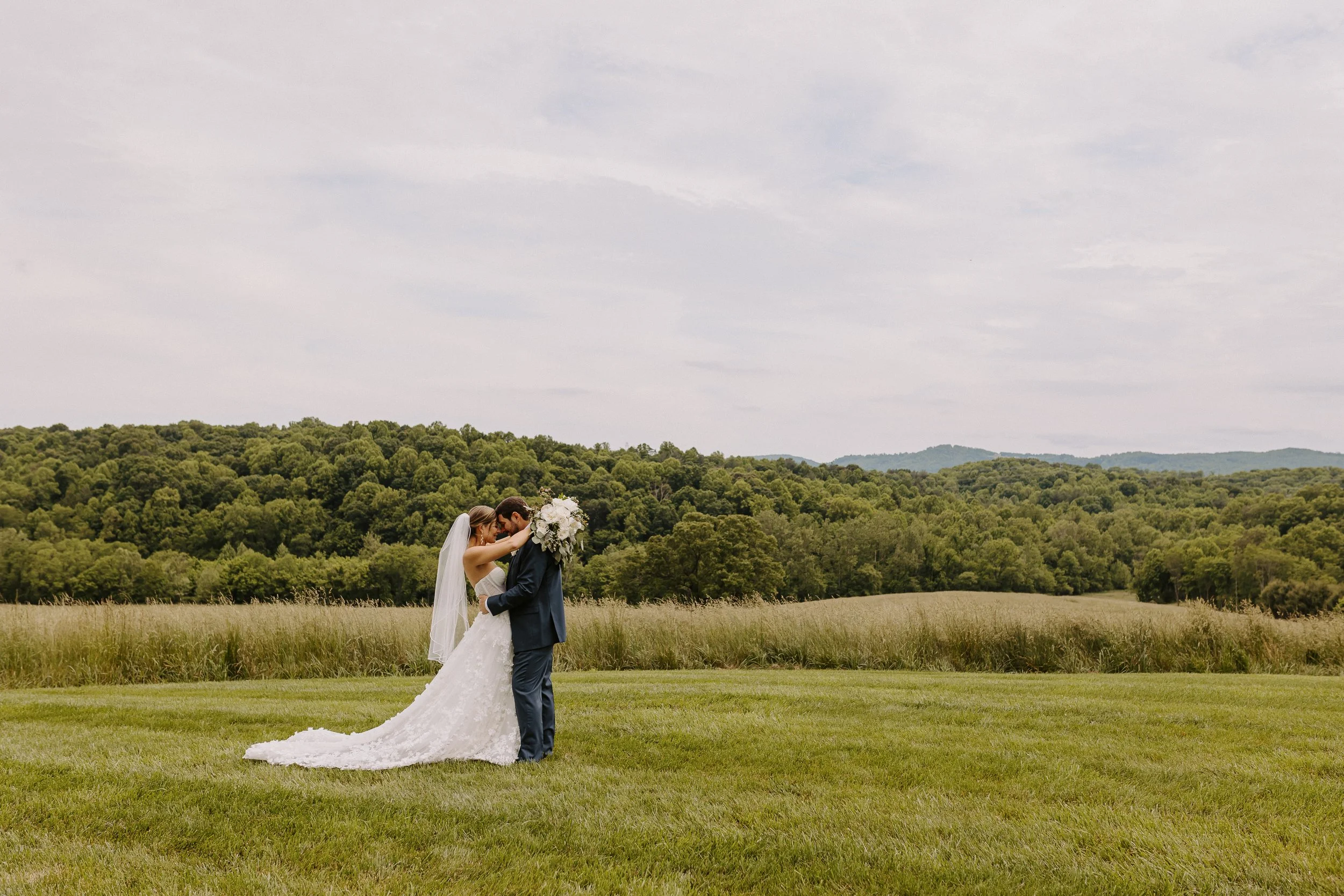 Bride and groom sharing a kiss outdoors on a grassy field with rolling hills and trees in the background.
