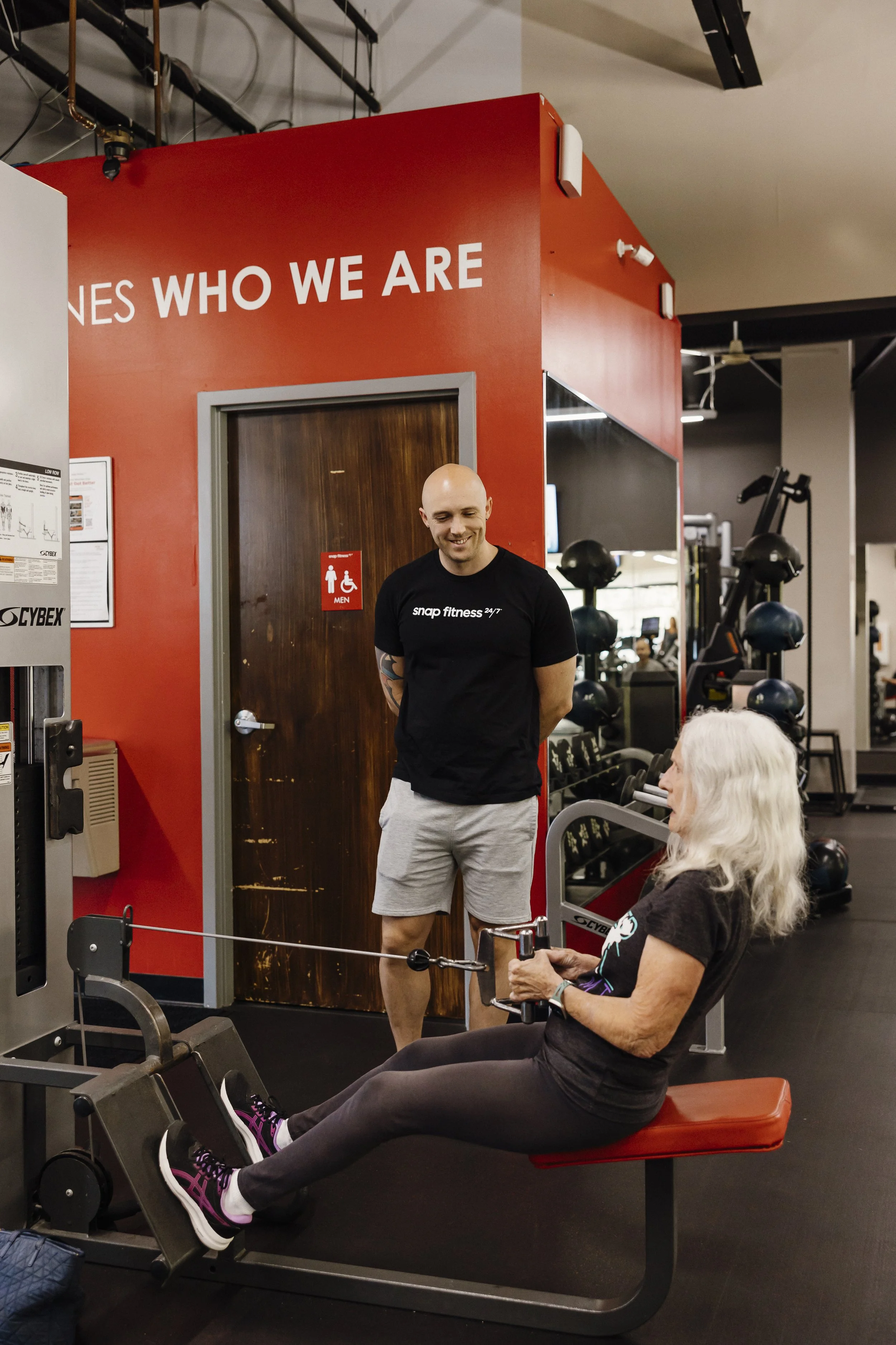 An elderly woman with gray hair exercising on a leg curl machine at a gym, with a male trainer standing nearby and smiling.