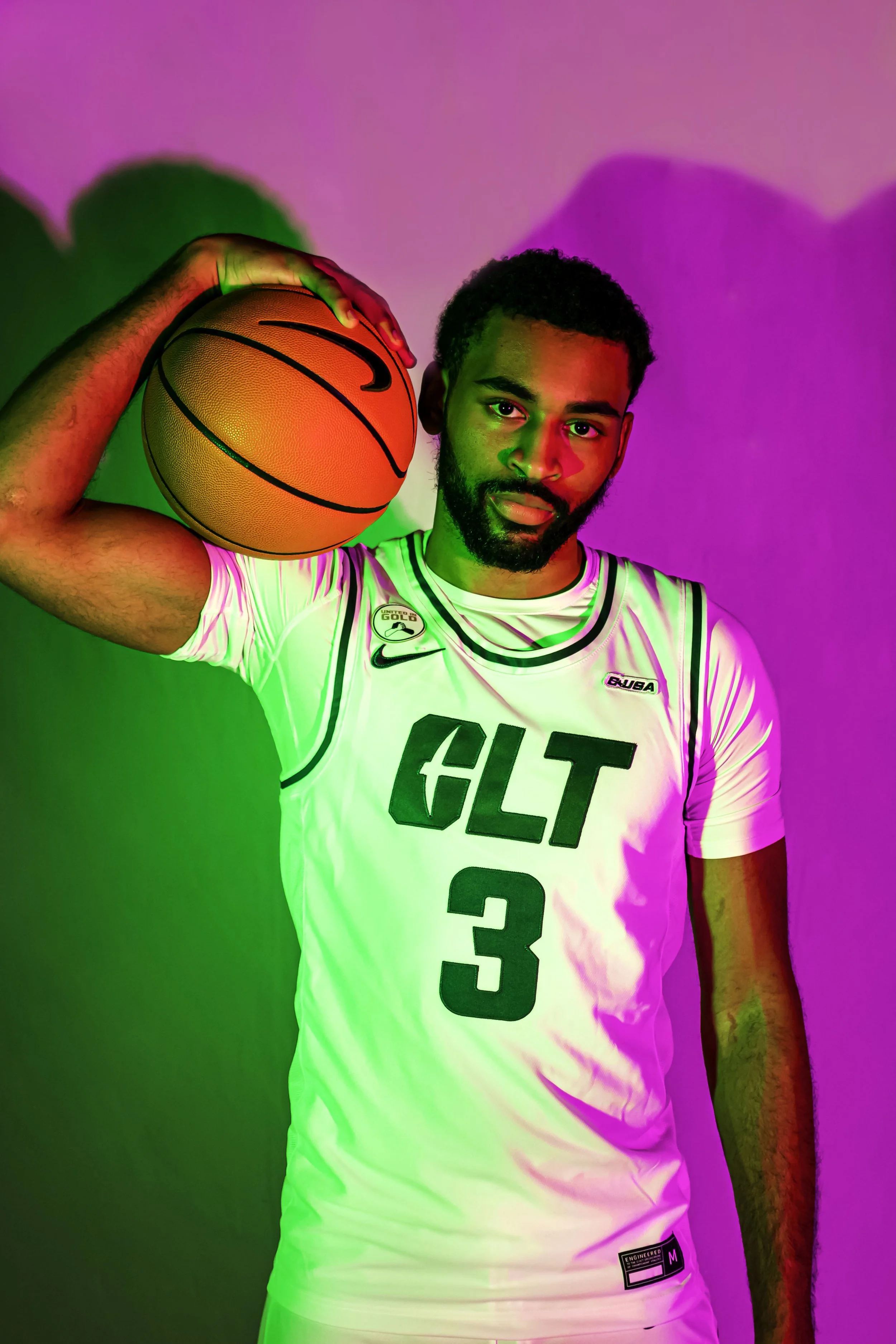 A young man with a beard and short curly hair posing with a basketball on his shoulder, wearing a white basketball jersey with black lettering 'CLT 3' and logos, against a colorful pink and green background.