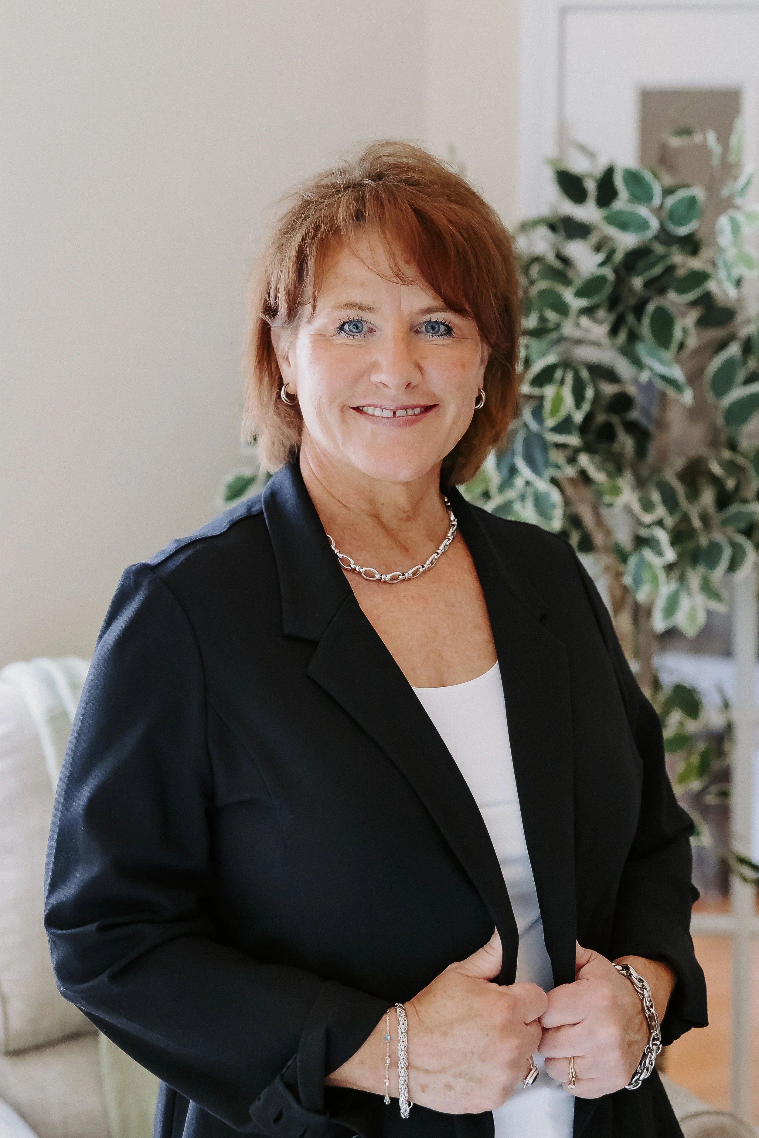 A smiling woman with red hair and blue eyes, wearing a black blazer, white top, and silver jewelry, standing indoors with a leafy plant in the background.