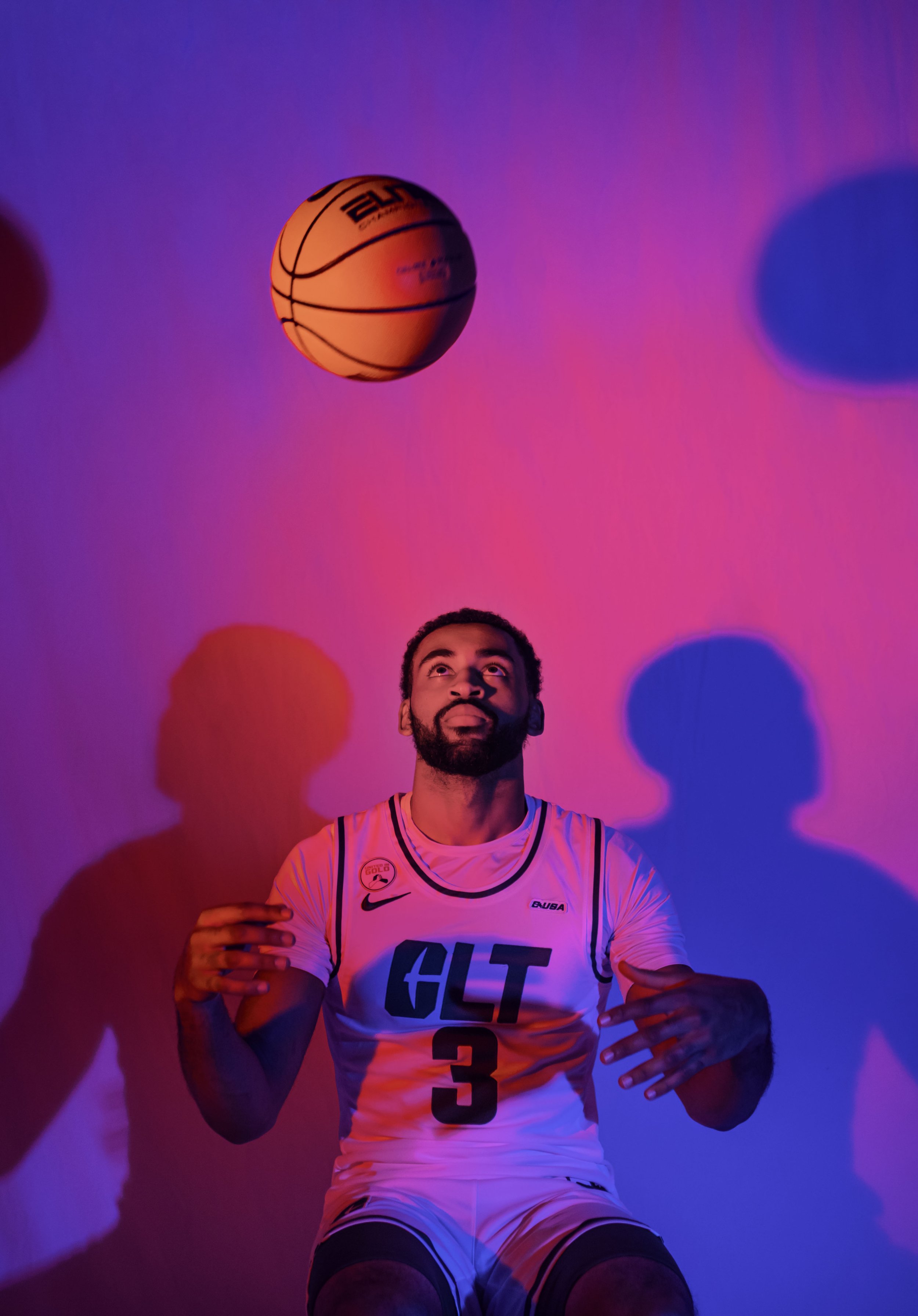 A basketball player sitting on the floor wearing a white jersey with the word 'GLT' and the number 3, looking up at a basketball in the air, with colorful pink and blue lighting creating shadows on the wall.